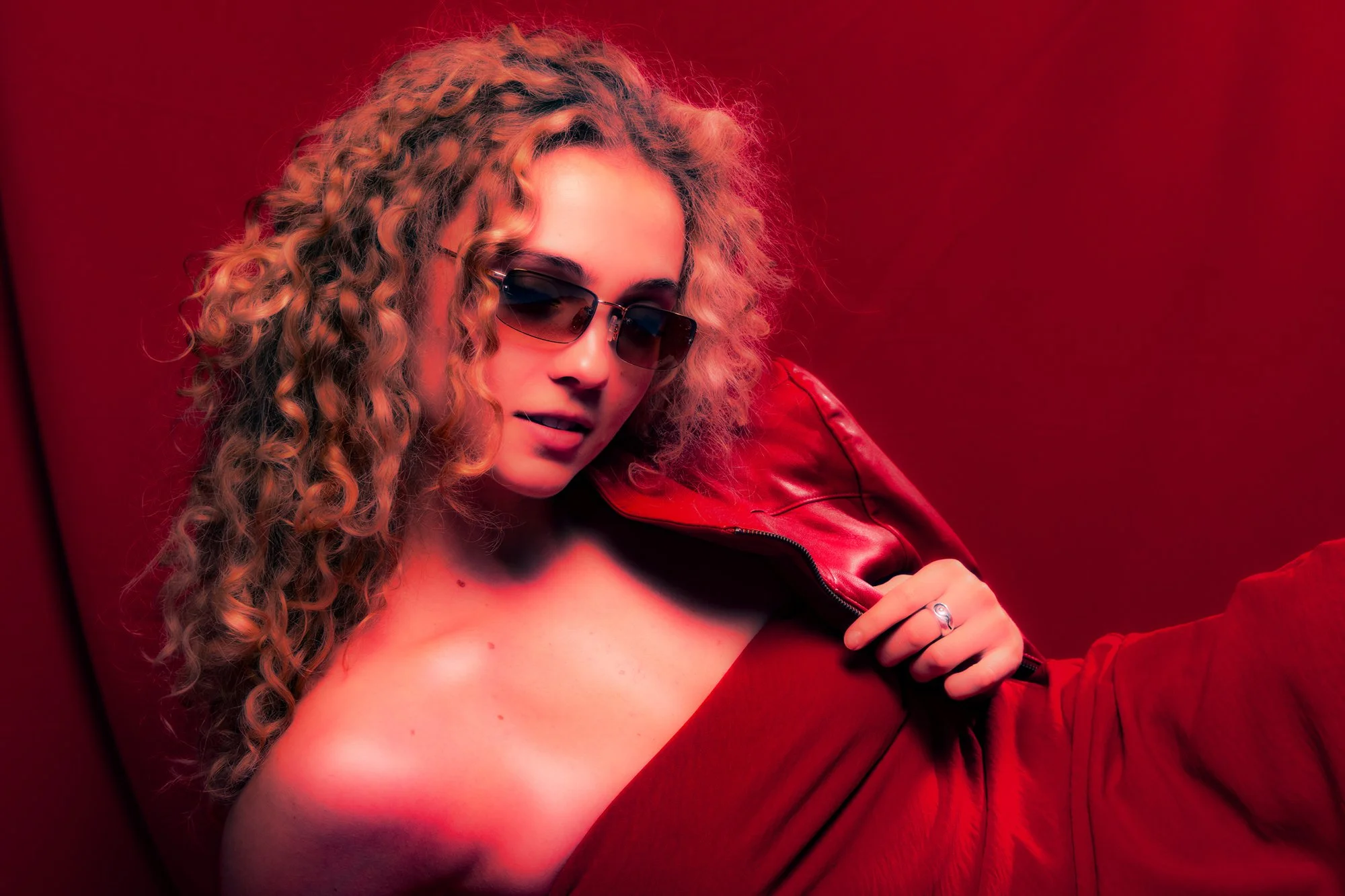 Woman with curly blonde hair wearing red tube dress lying across frame with sunglasses on, holding red leather jacket over shoulder against red backdrop, strong red tones.