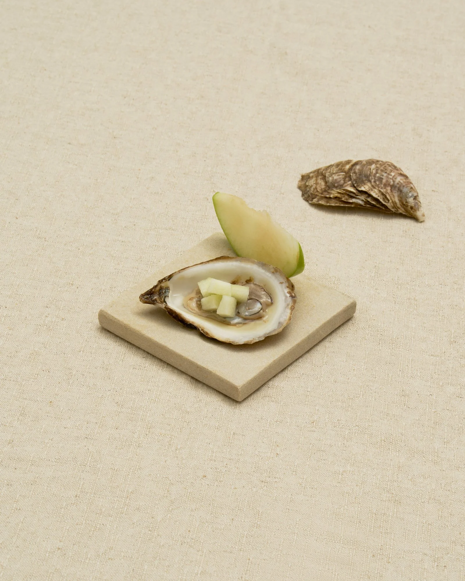 A serving of fresh oysters topped with diced cucumber, accompanied by a slice of green apple and an unopened oyster shell on a textured beige surface.