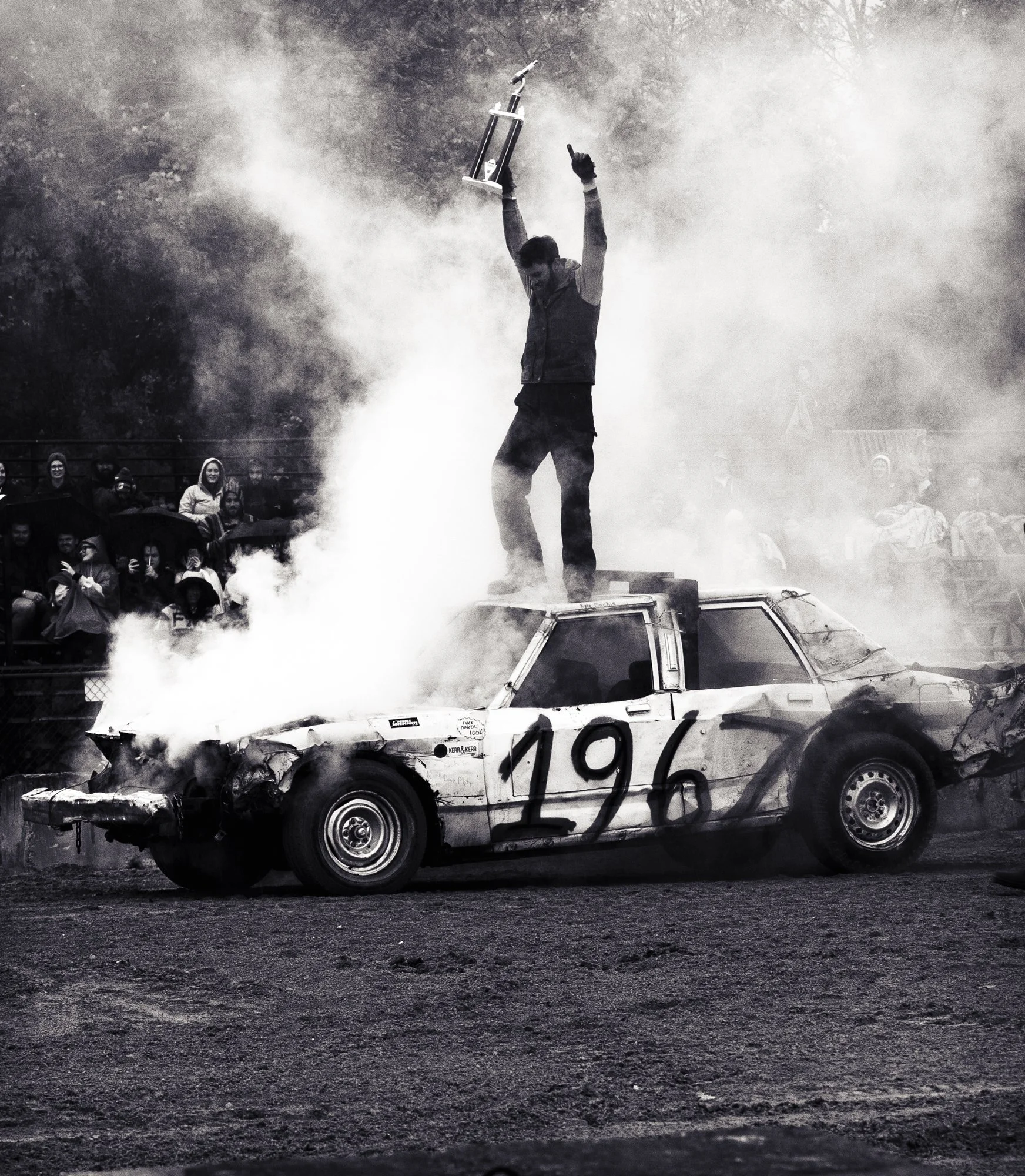 Man standing on top of damaged smoking car holding trophy, grainy black and white, audience behind