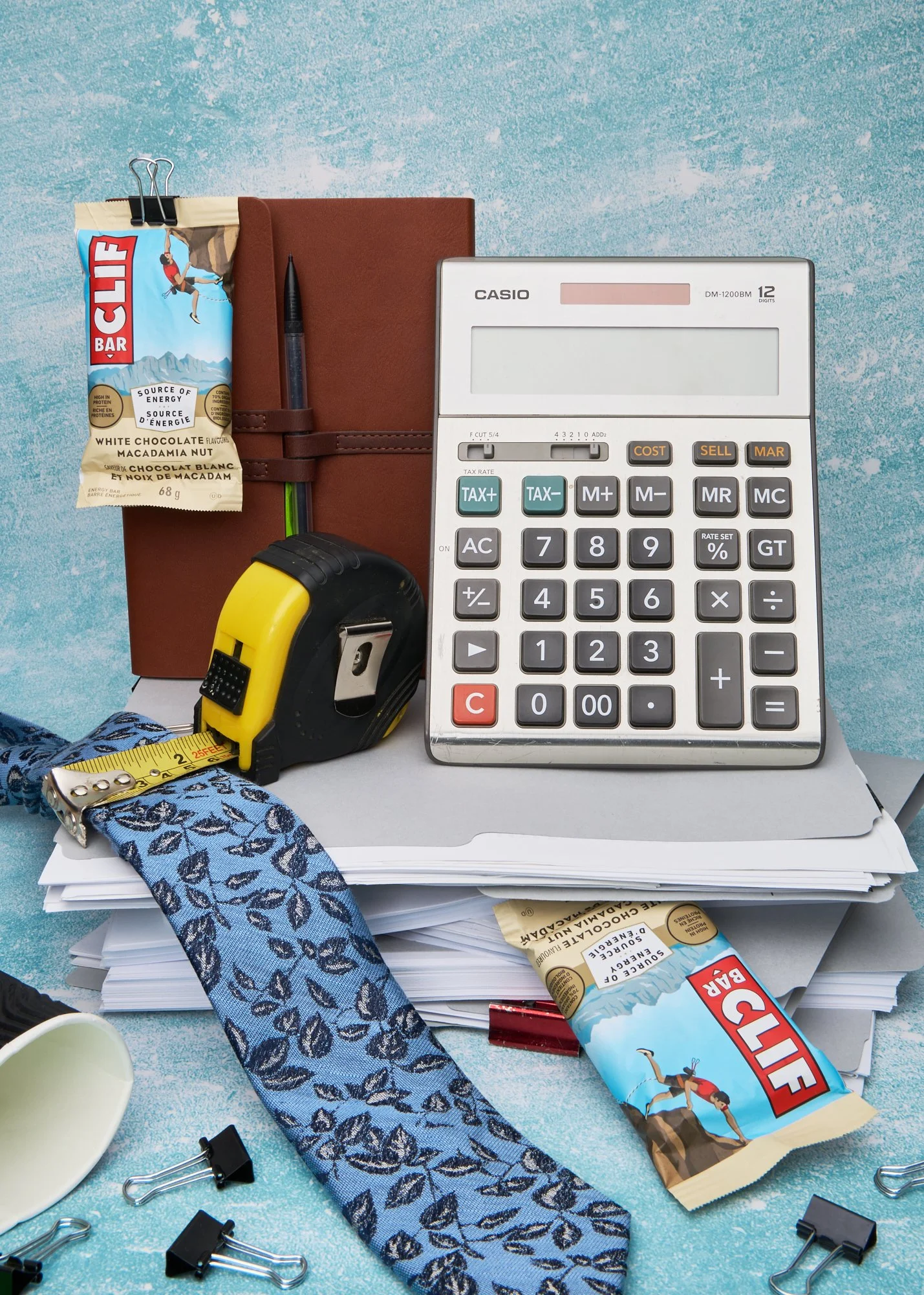 Office still life with calculator, notebook, tape measure on stacked papers with Clif bar and blue patterned tie.