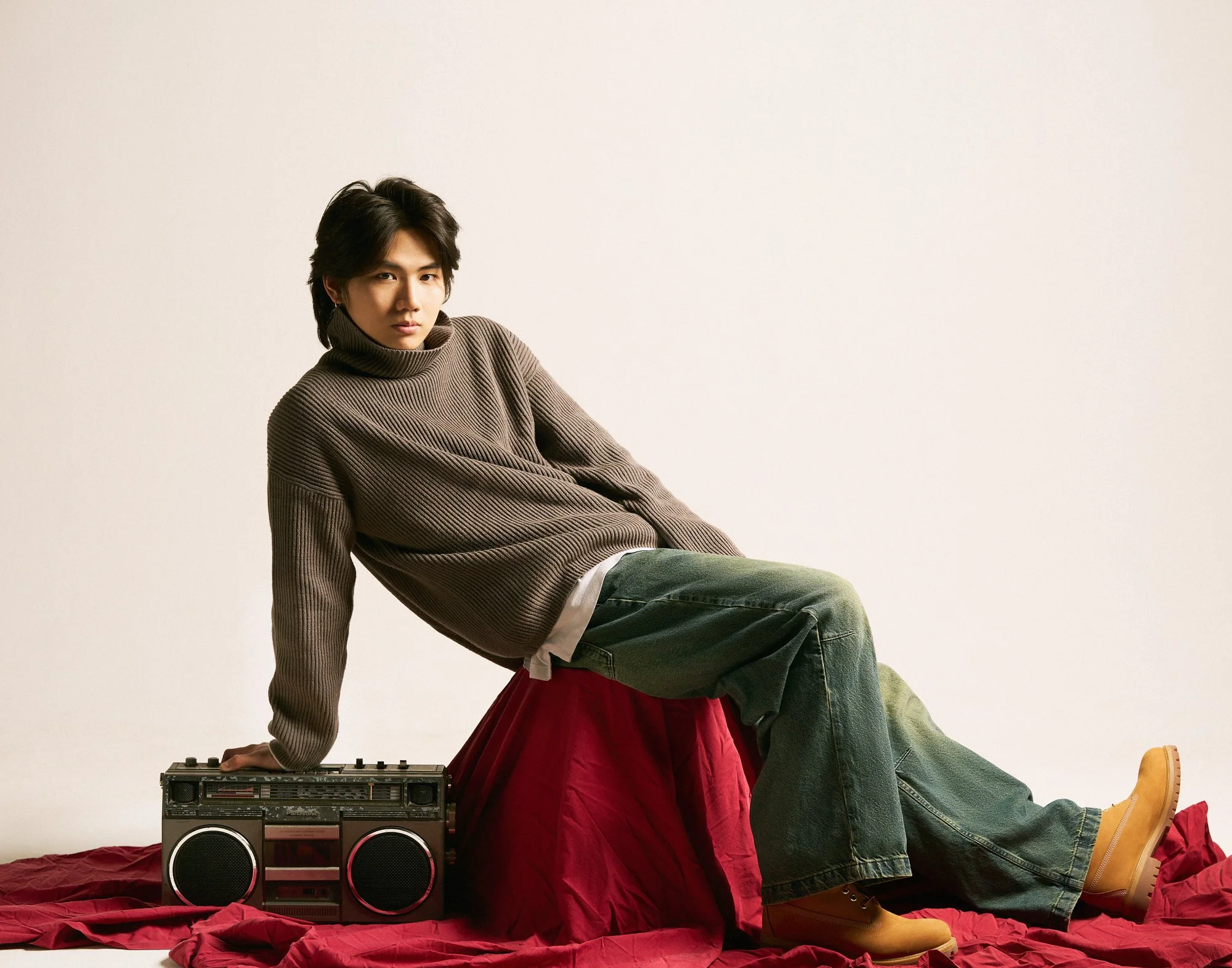 Young man with dark hair sits on a red fabric-covered surface, wearing a brown turtleneck sweater, blue and yellow jeans, and yellow boots, with a vintage boombox nearby, against a plain light background.