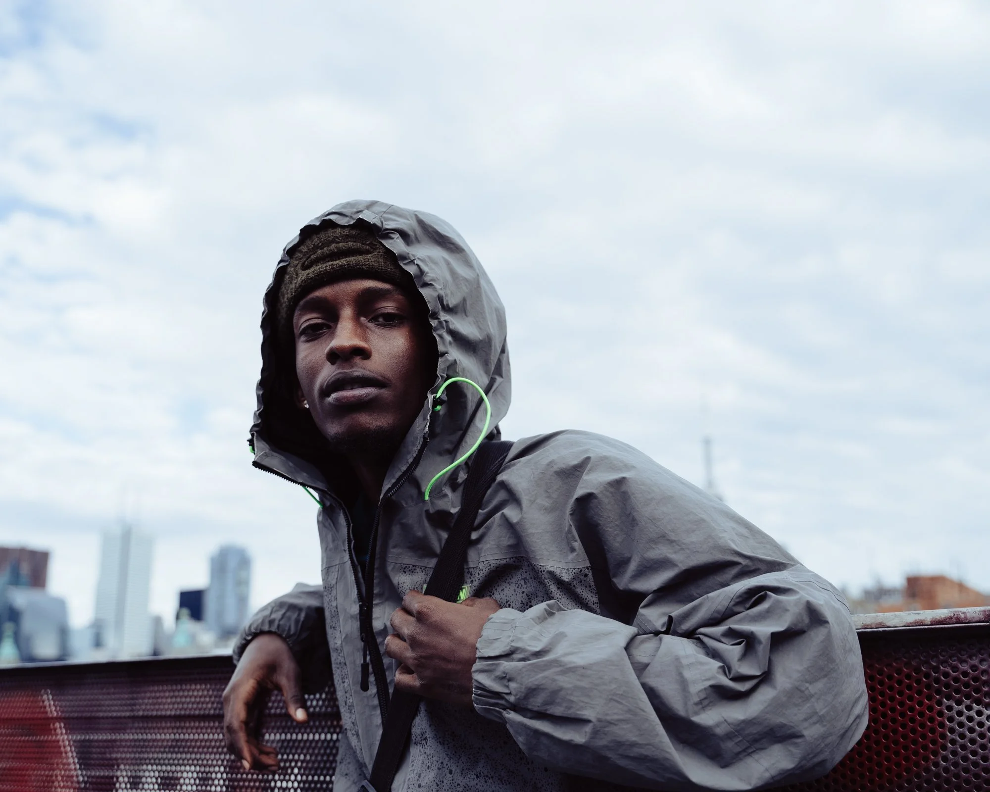 Man wearing windbreaker, hood up, looking at camera, clutching shoulder strap, leaning on rail, sky background.