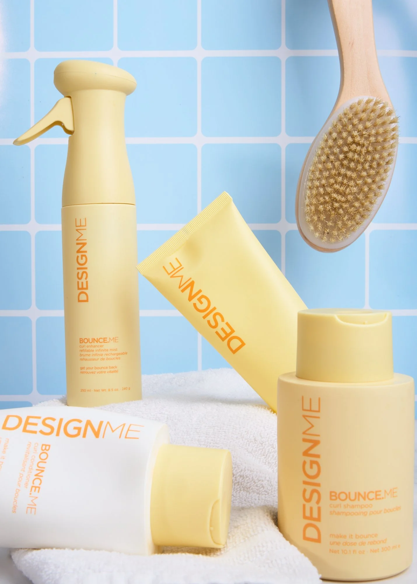 Yellow DesignMe shampoo, conditioner, and curly hair products on white towel, wooden brush hanging against blue tiled background.