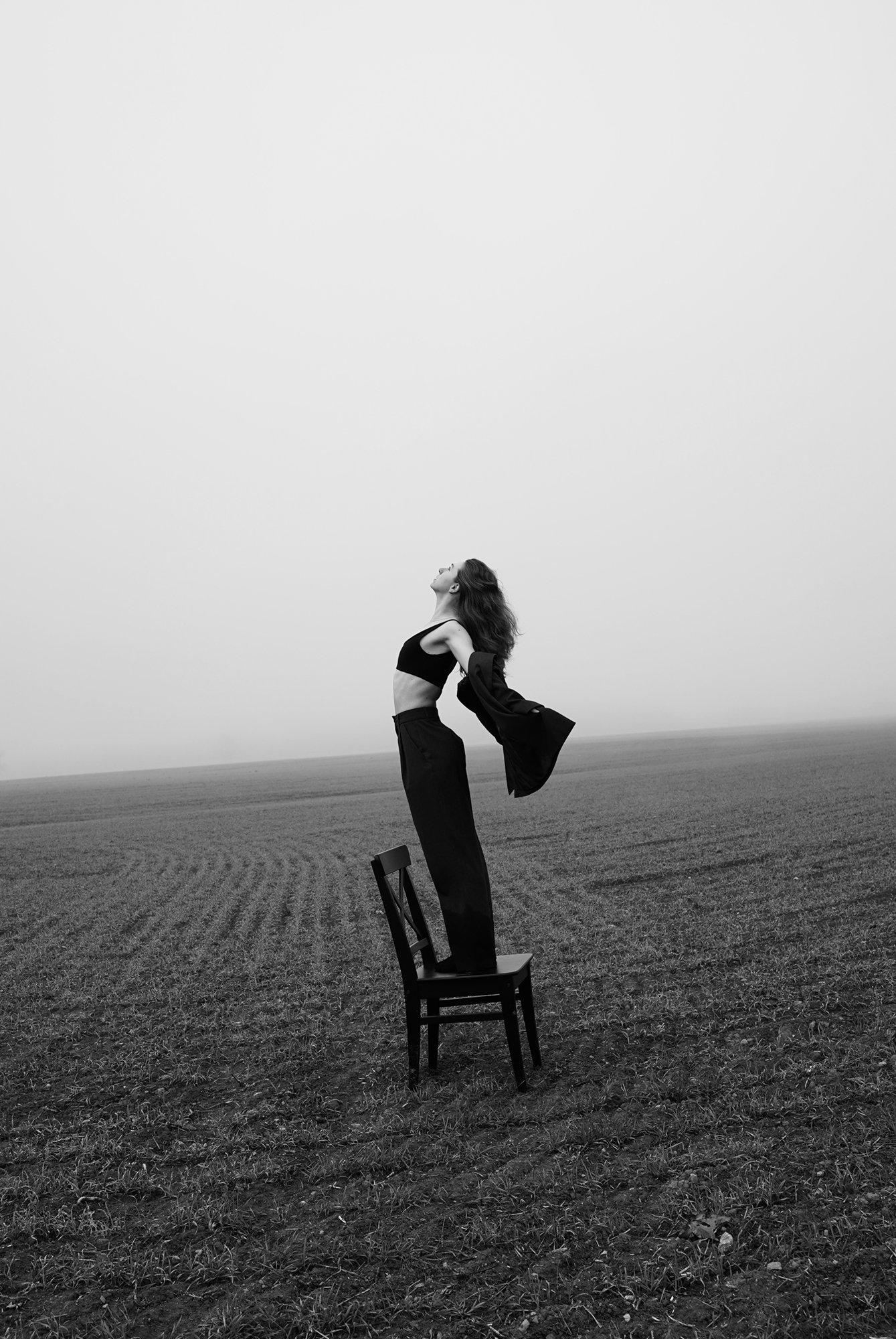 Woman in centre of frame standing on chair in foggy field looking up at sky, wearing dress pants, bra top and pushing out blazer with arms. Black and white tilted horizon line.