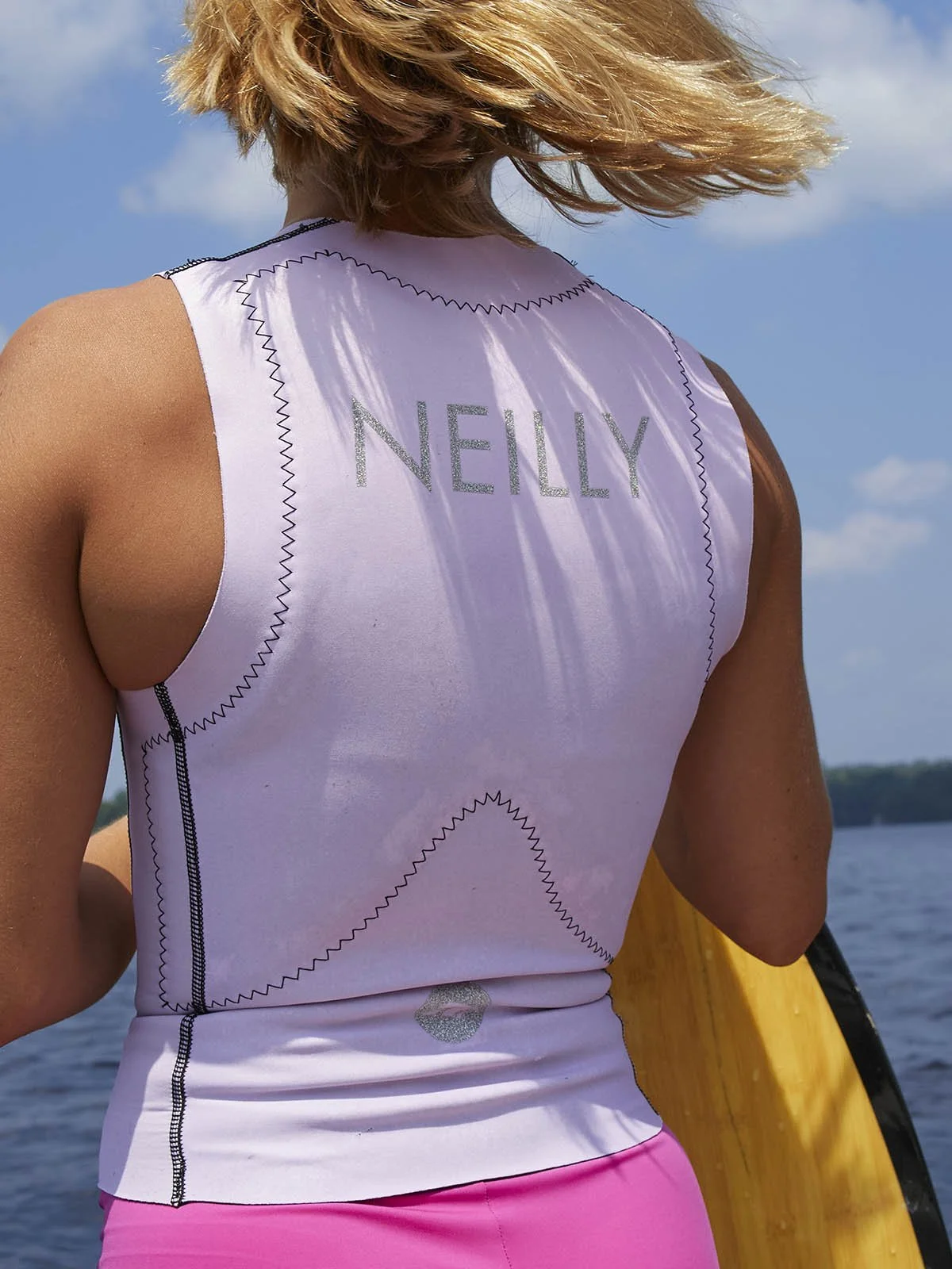 Woman in studded lifejacket facing away from camera holding surfboard with hair swaying in wind on sunny day