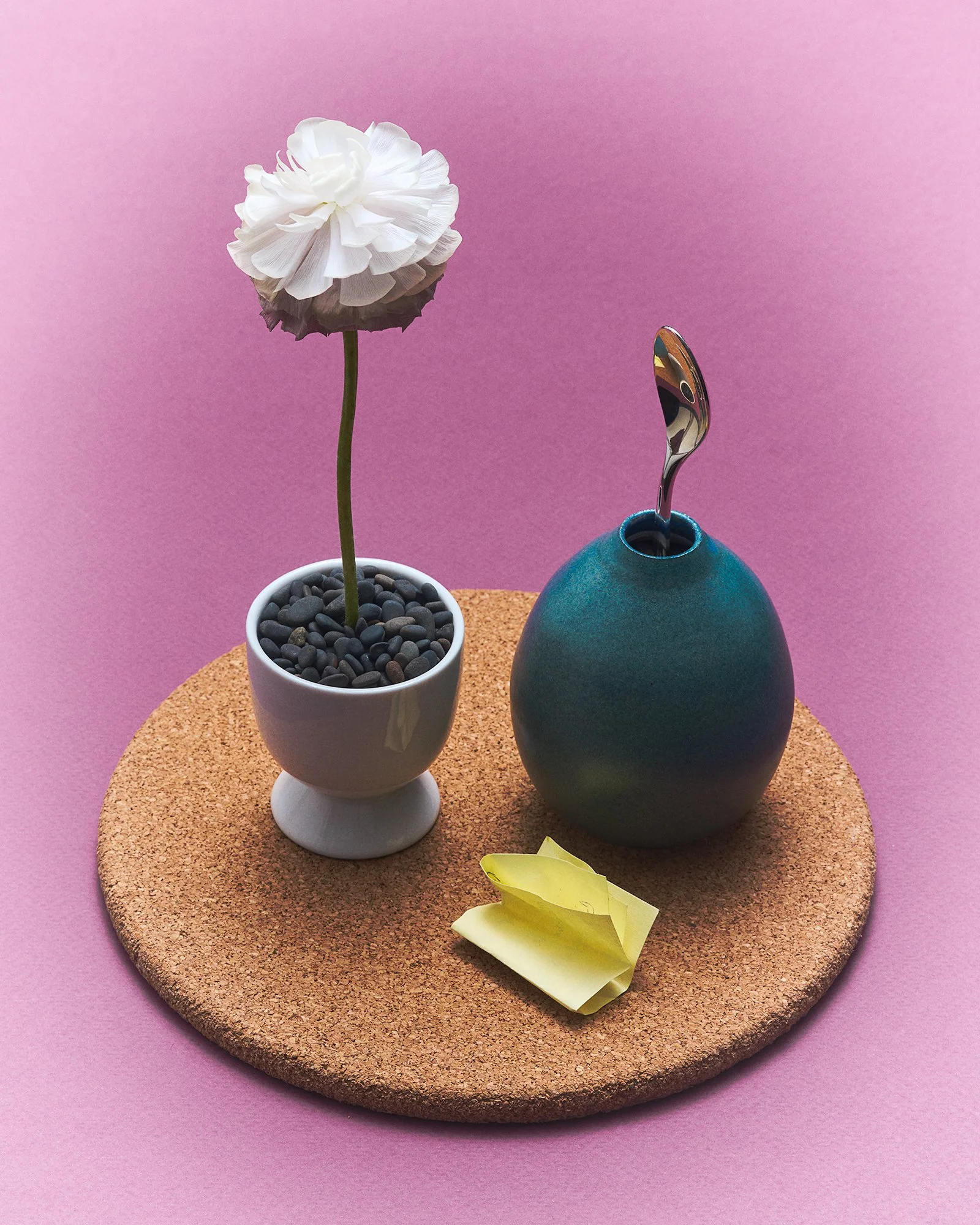 Still-life with white flower in small white pot, beside matte blue vase holding small upright spoon, folded yellow paper note, on round cork surface against pink background.