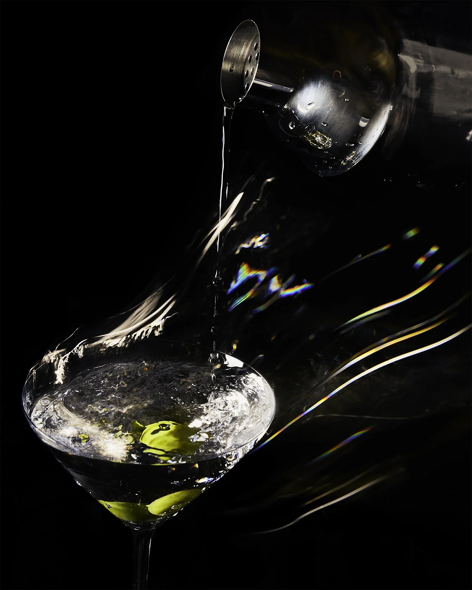 Martini photograph showing liquid being poured, olive garnish, with dragged shutter and strobe lighting. Motion blur and prismatic reflections against dark background.