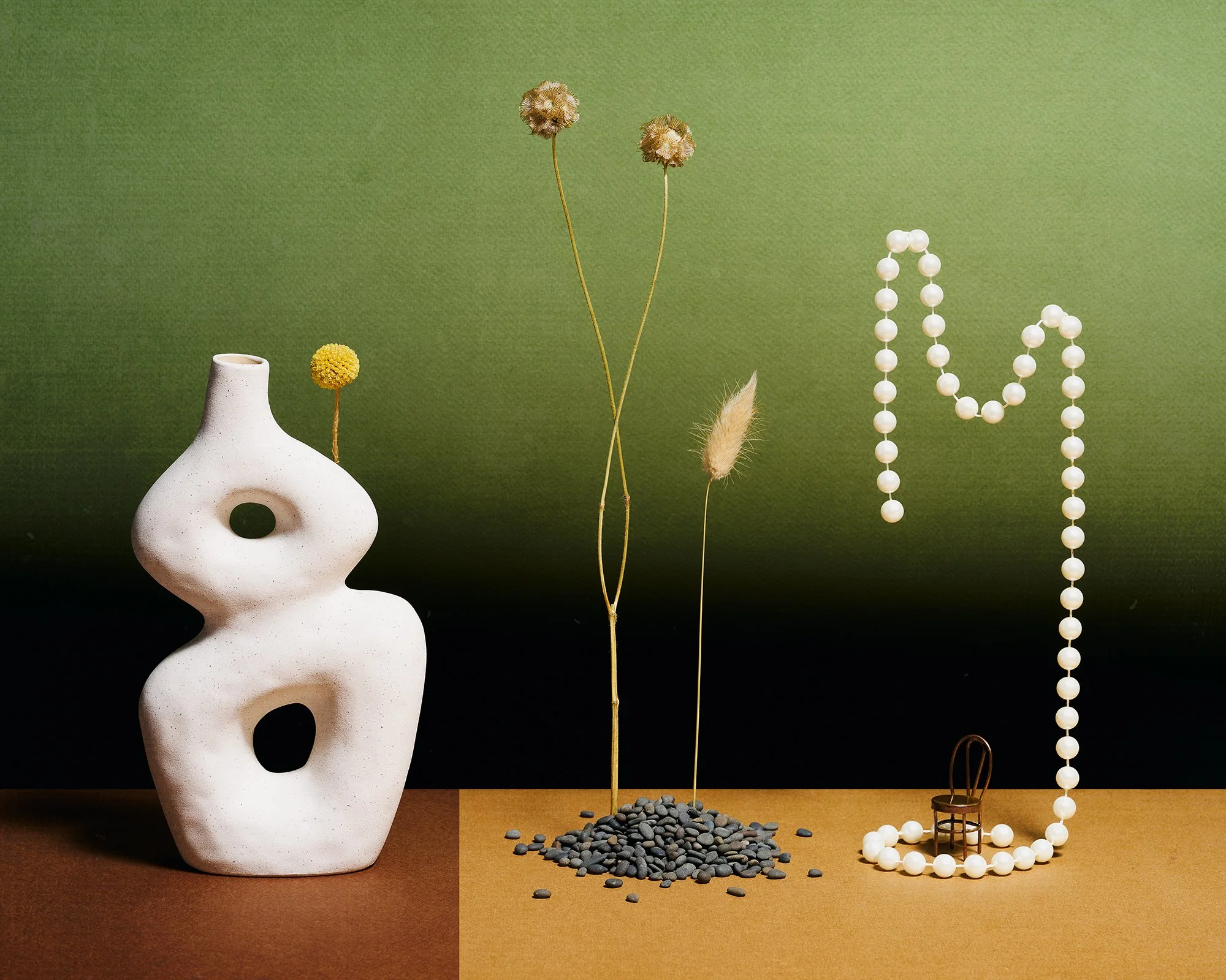 Still-life with white vase, small pile of black stones and dried stems dried stems, floating string of pearls around tiny chair on brown surface and green ombre background.