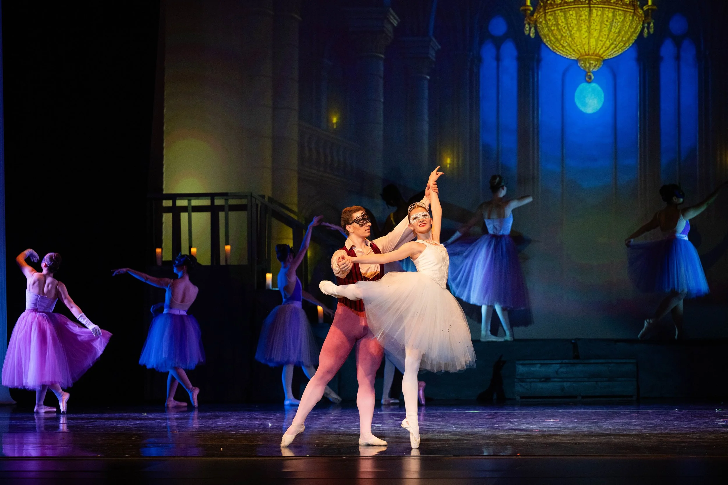 Ballet dancers performing on stage with a moonlit backdrop and elegant chandeliers. The focus is on a male and female dancer in the center, dressed in classical ballet costumes, with other ballerinas in tutus dancing in the background.