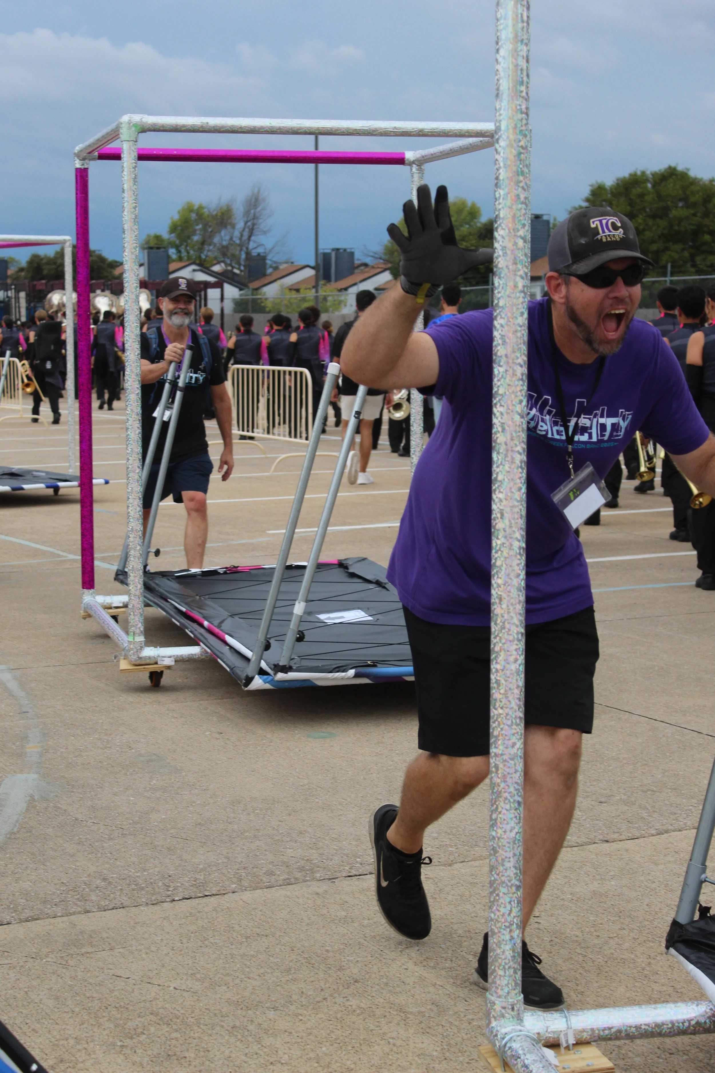 A man wearing a purple T-shirt, black shorts, a cap, and sunglasses runs through a finish gate at an outdoor event, excited and smiling, with a large crowd of people in the background.