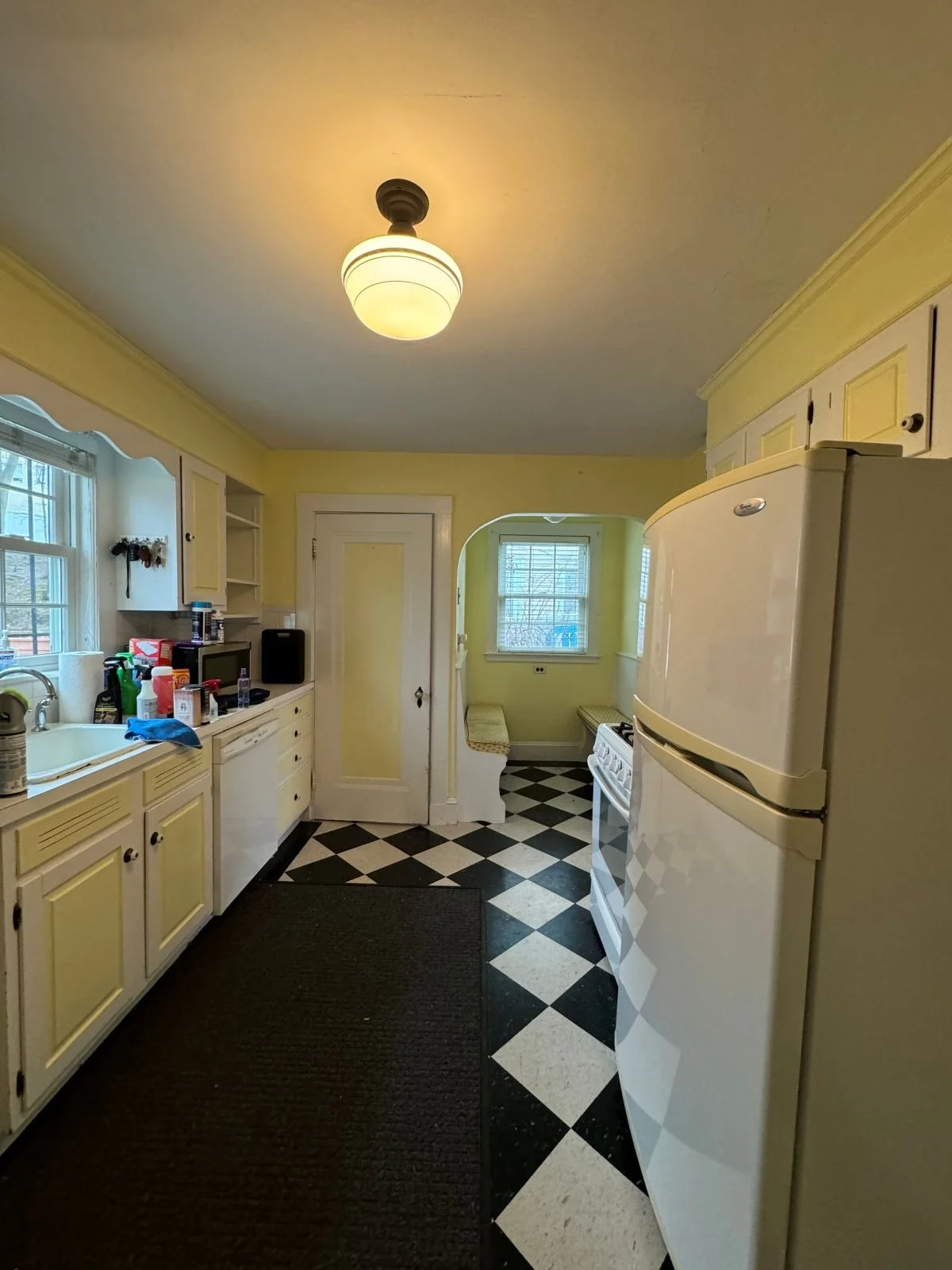 Existing kitchen overview from living room looking at outdated refrigerator, black and white linoleum floors, two-toned yellow and white cabinetry with a single surface mount light fixture in the middle of the room