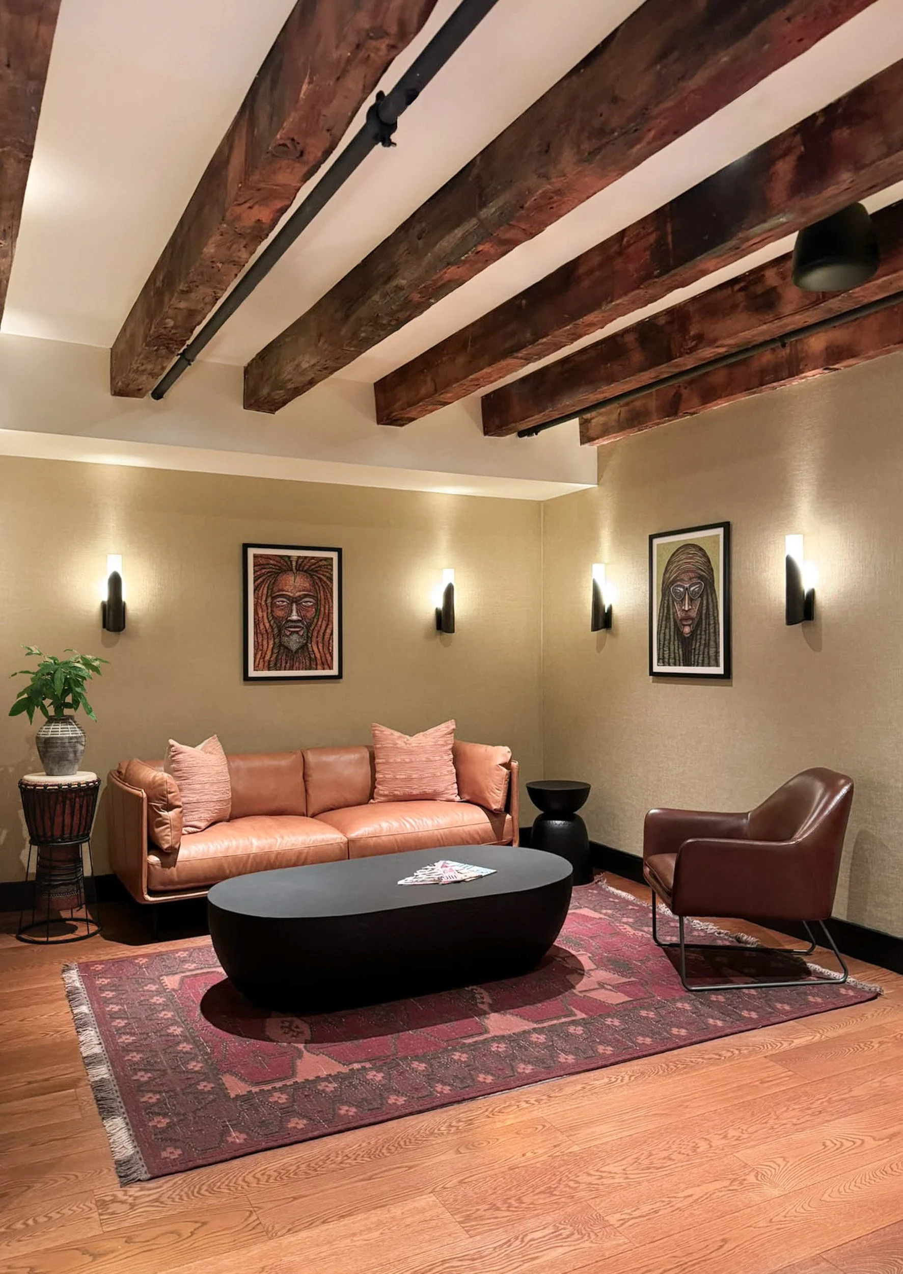 Retail seating area with custom Jamaican art, leather couch and chair, black accent pieces over red rug with black sconce lighting under original heavy timber beams.