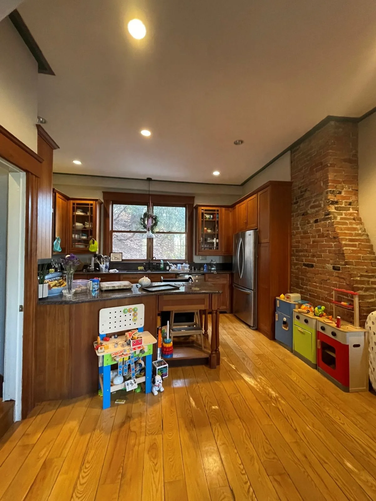 Dark 90's kitchen with stained wood cabinetry, a brick chimney stack, and limited natural light prior to architectural reconfiguration.