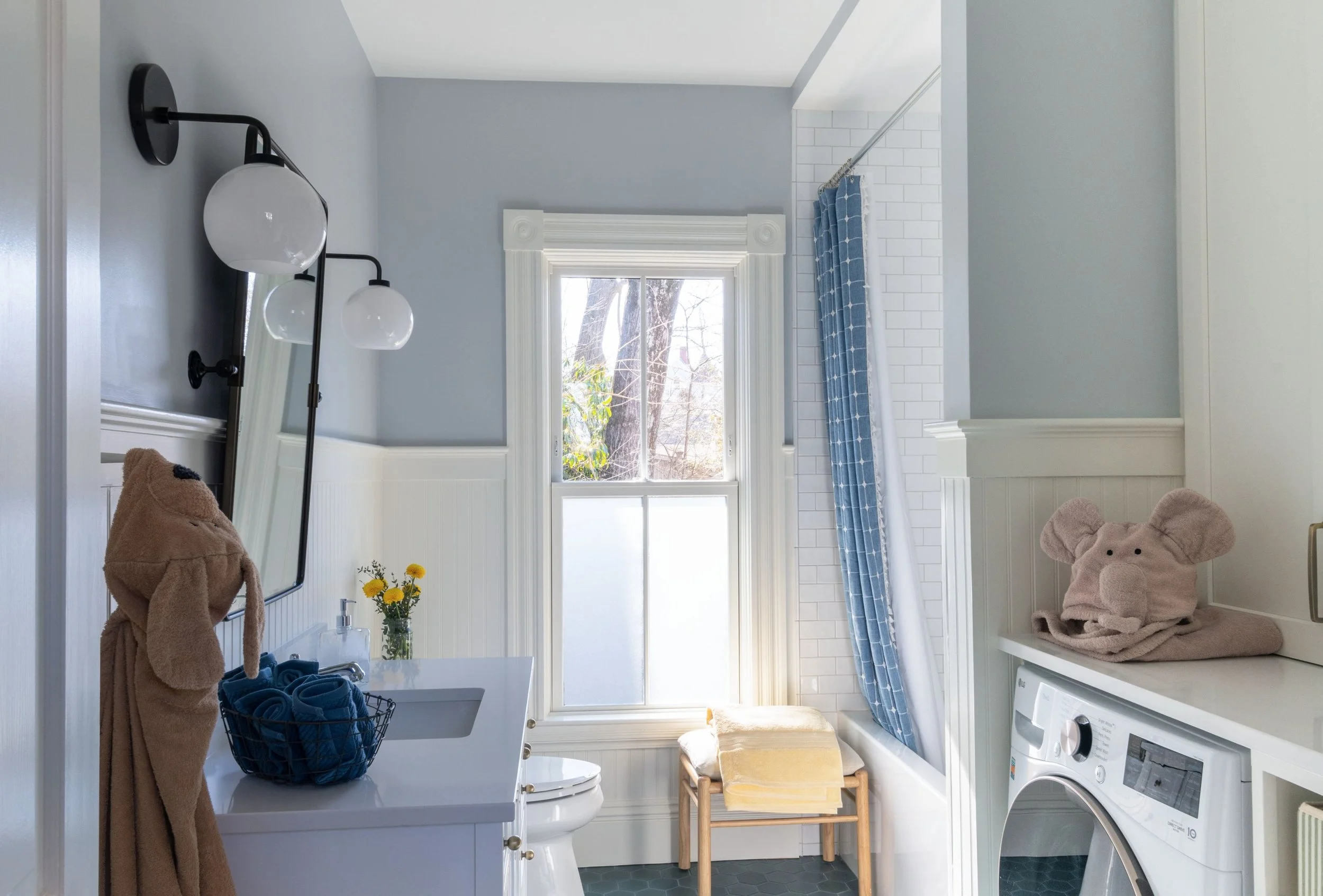 Renovated kids bathroom and laundry of an 1846 Victorian home in Newton MA featuring new window with refurbished casing, teal hexagon floor tile, integrated washer, white vanity, wainscoting, matte black sconce and subway tile tub surround