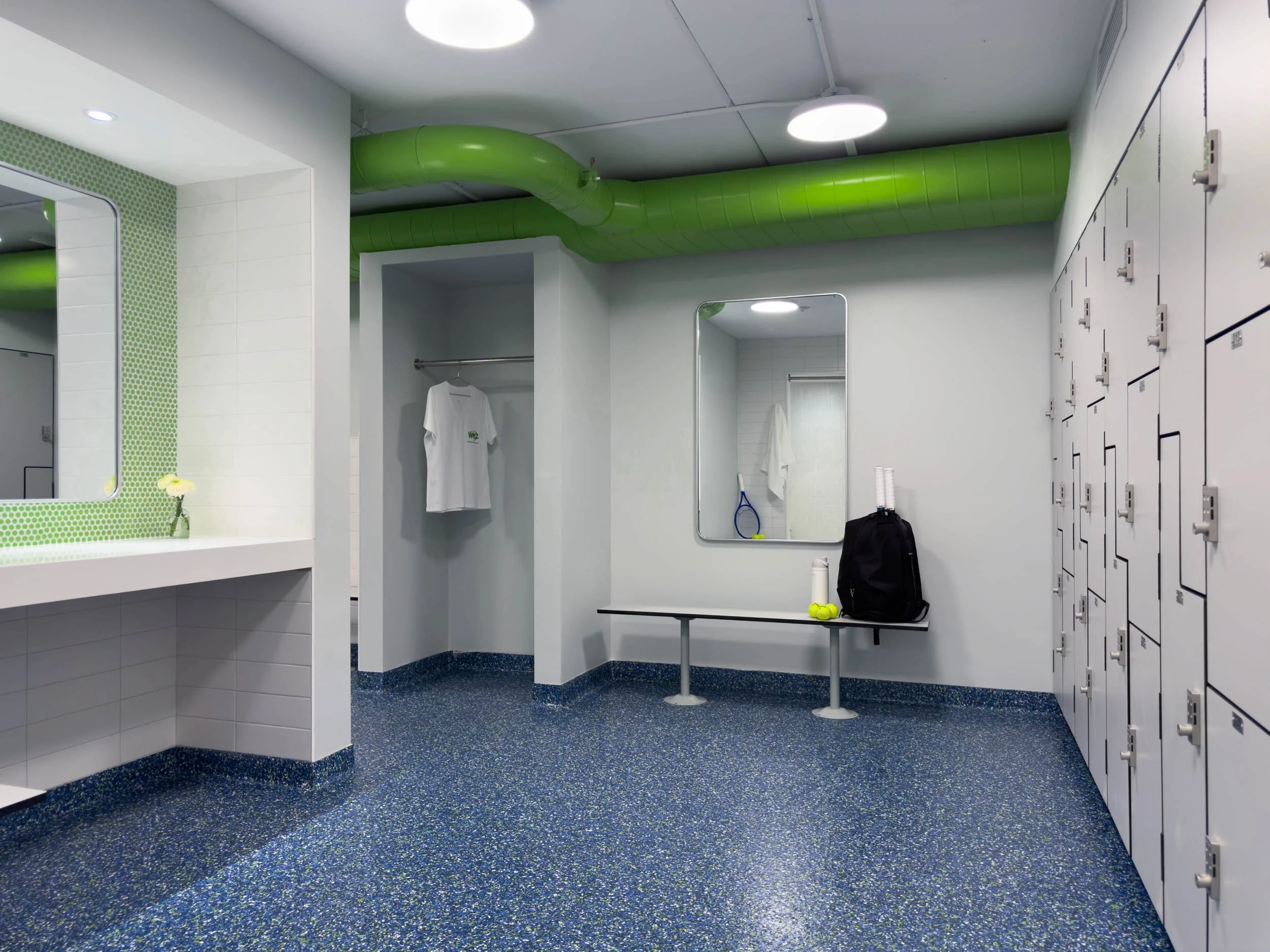 women's locker room changing area with epoxy floors, vanity counter that has green penny tile backsplash and exposed green ductwork  with grey lockers.