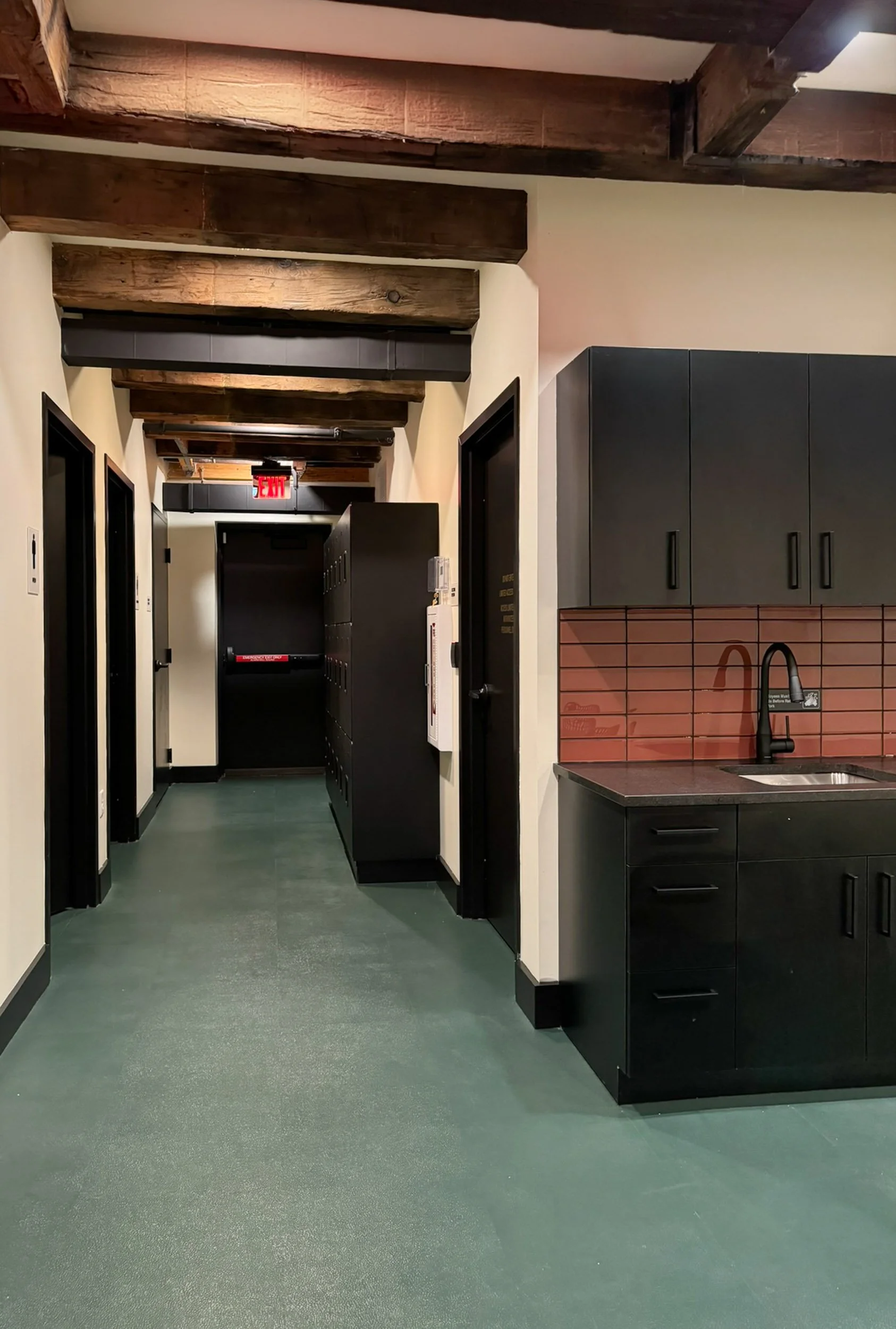 A hallway leading to an employee break room kitchenette with black cabinetry, terracotta glass tile backsplash, a sink and exposed wood beams.