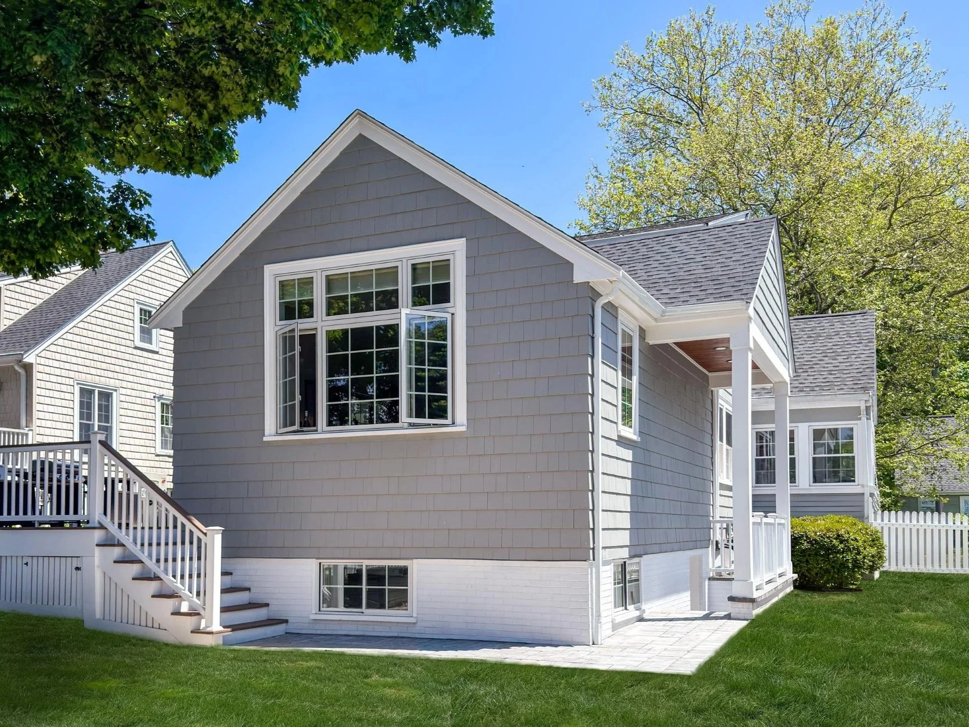 Side angle view of a completed vaulted kitchen addition on a colonial home in Melrose, MA showing the new roofline, picture windows, basement stair walkout, and paver patio, designed by Tact and Grit Design Co.