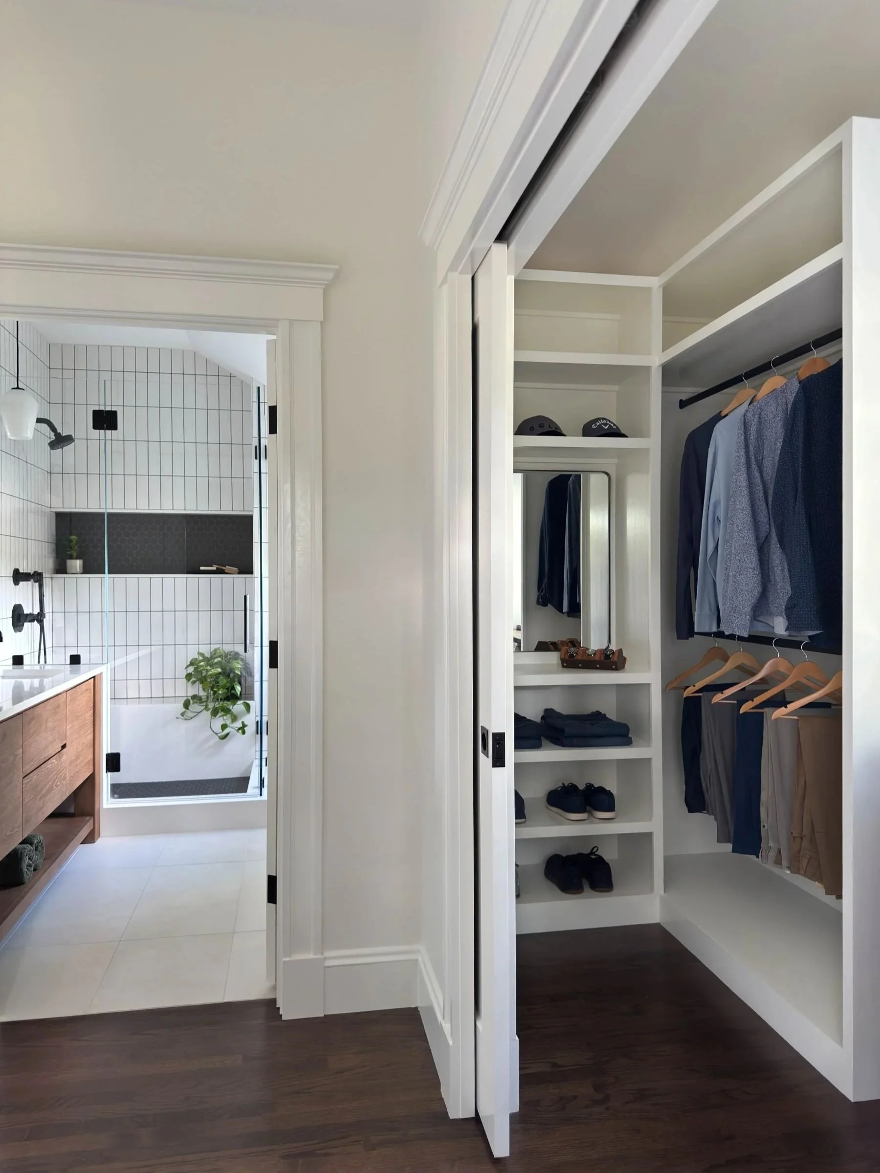 His side of a his and hers walk-in closet in a Dutch Colonial primary suite addition in Greater Boston viewed through pocket door showing custom built-in shelving with view beyond to primary bathroom designed by Tact and Grit Design Co.