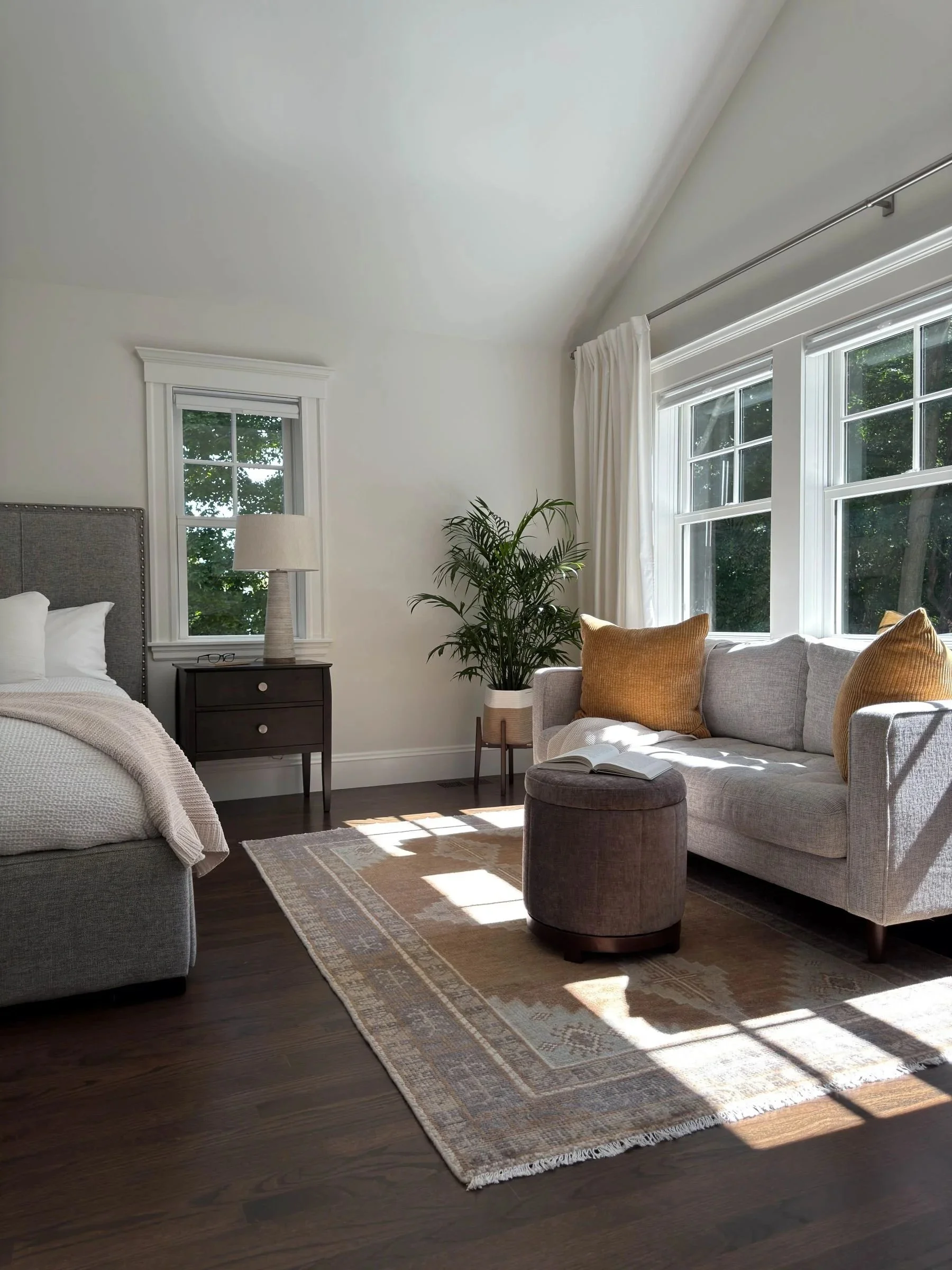Completed primary suite bedroom of a Dutch Colonial second story addition in Greater Boston showing reading area, vaulted ceiling and triple window array with natural light designed by Tact and Grit Design Co.