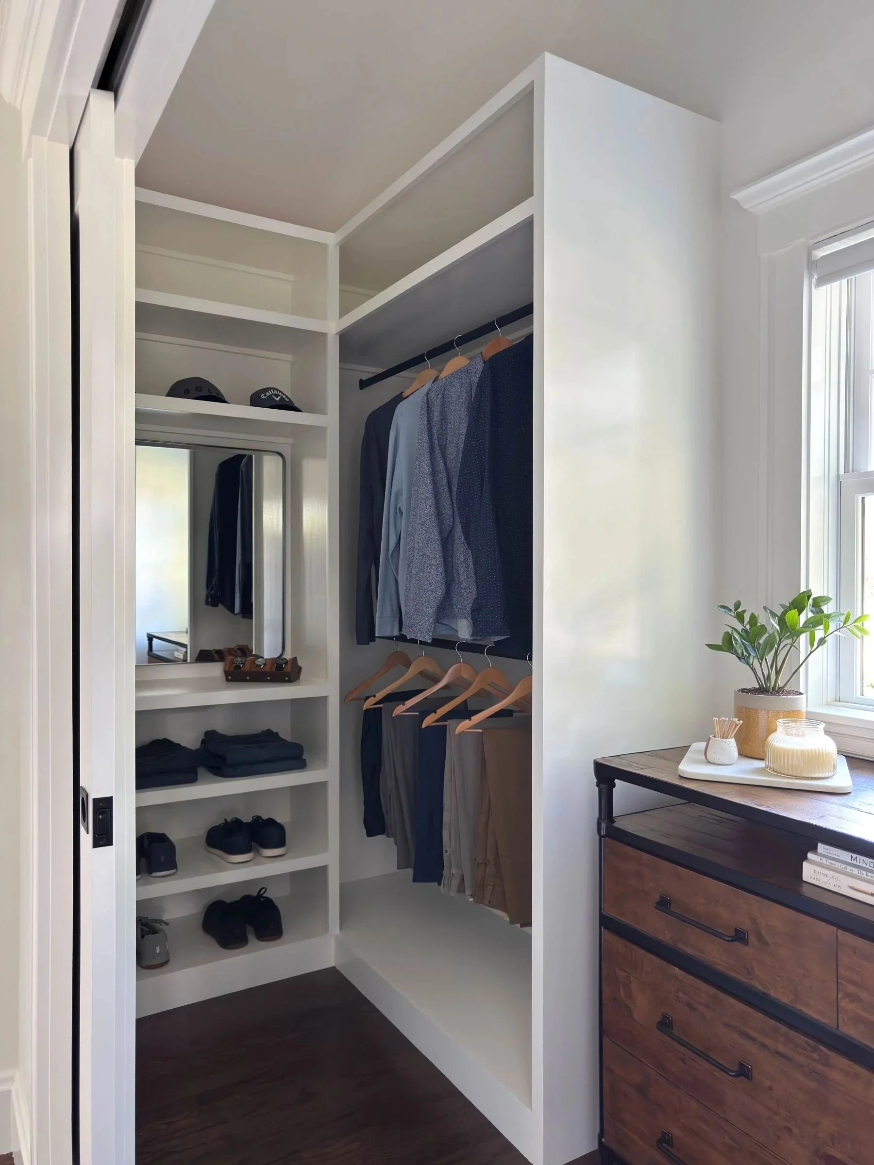 Completed walk-in closet of a Dutch Colonial primary suite addition in Greater Boston showing custom built-in shelving, hanging rods and reclaimed wood and iron dresser under window designed by Tact and Grit Design Co.