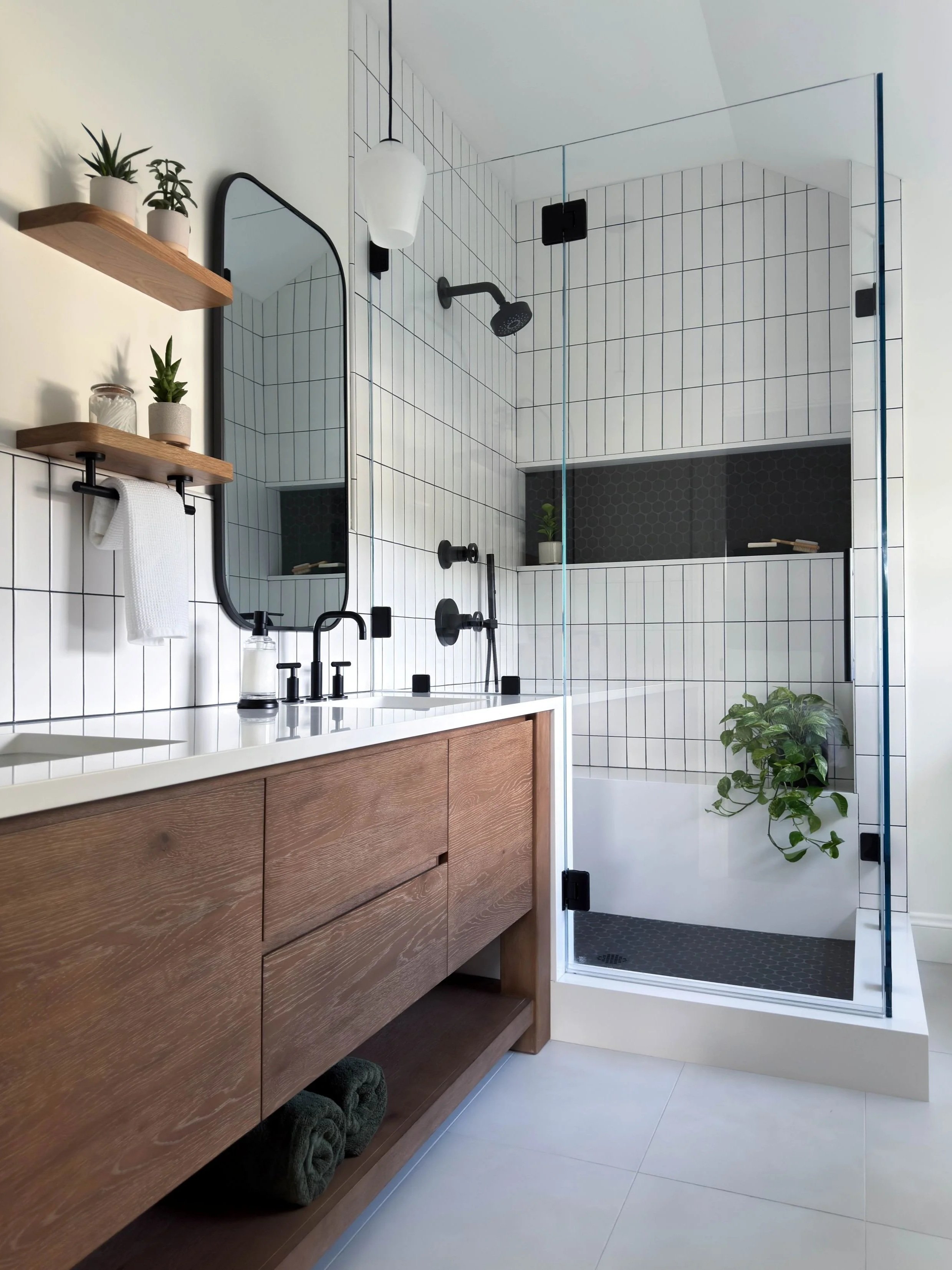 Double vanity features oak floating shelves with white quartz countertops sliding into shower and down to double mitered curb with clear glass enclosing a black and white modern shower with black circular fixtures