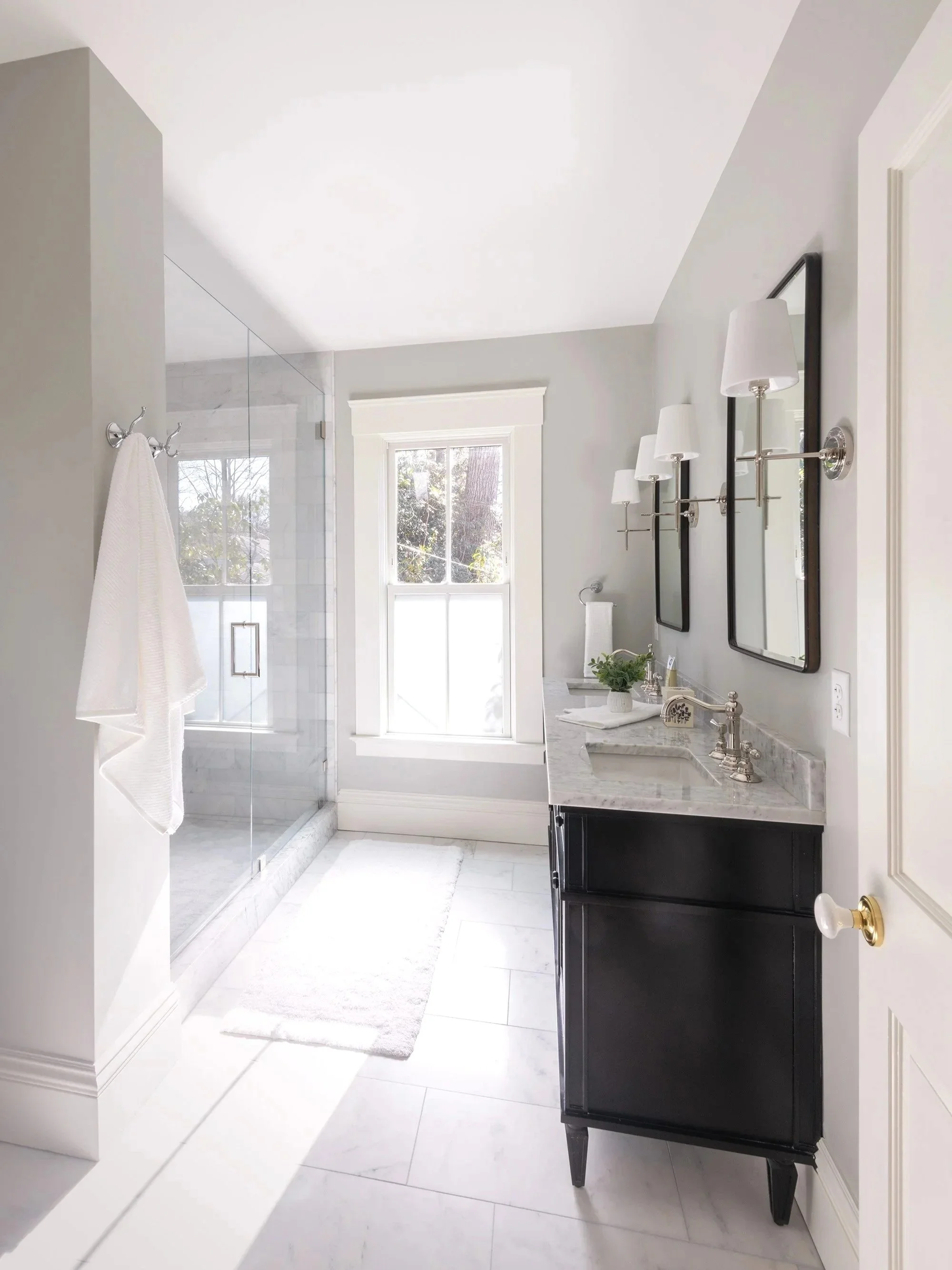 Renovated primary bathroom of an 1846 Victorian home in Newton MA featuring dark double vanity, marble tile and glass shower enclosure.