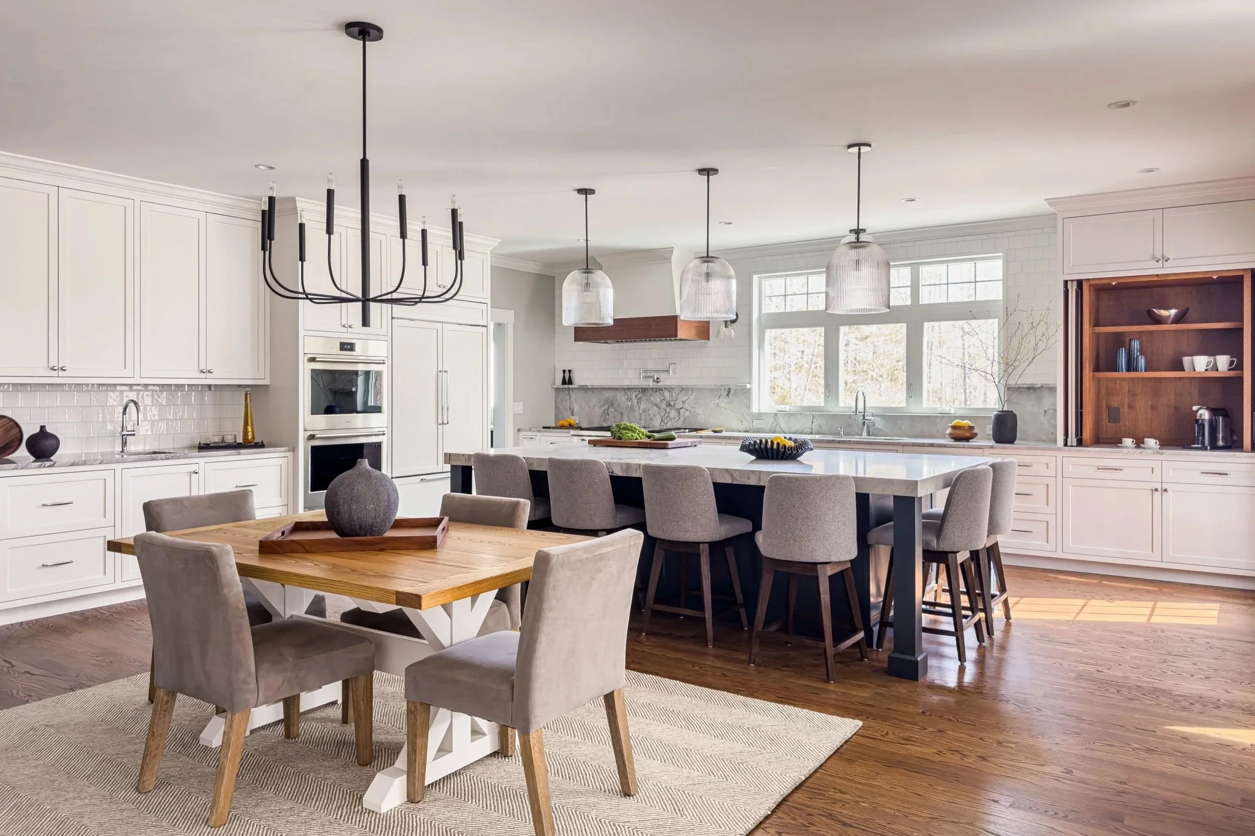 Dining area with four chairs and black chandelier with kitchen beyond with white cabinetry with quartzite countertops and large navy island with walnut stained oak coffee cabinet and range hood accents 