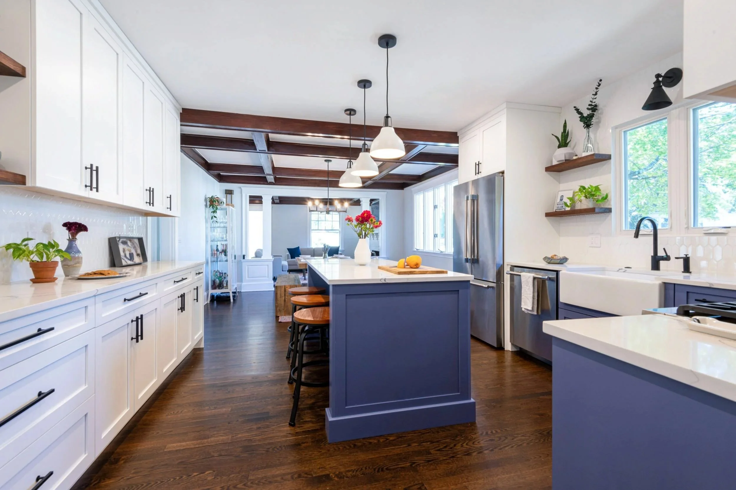 Expansive white kitchen with periwinkle blue accent cabinetry and black hardware with a rich brown coffered ceiling treatment in the open dining room beyond.