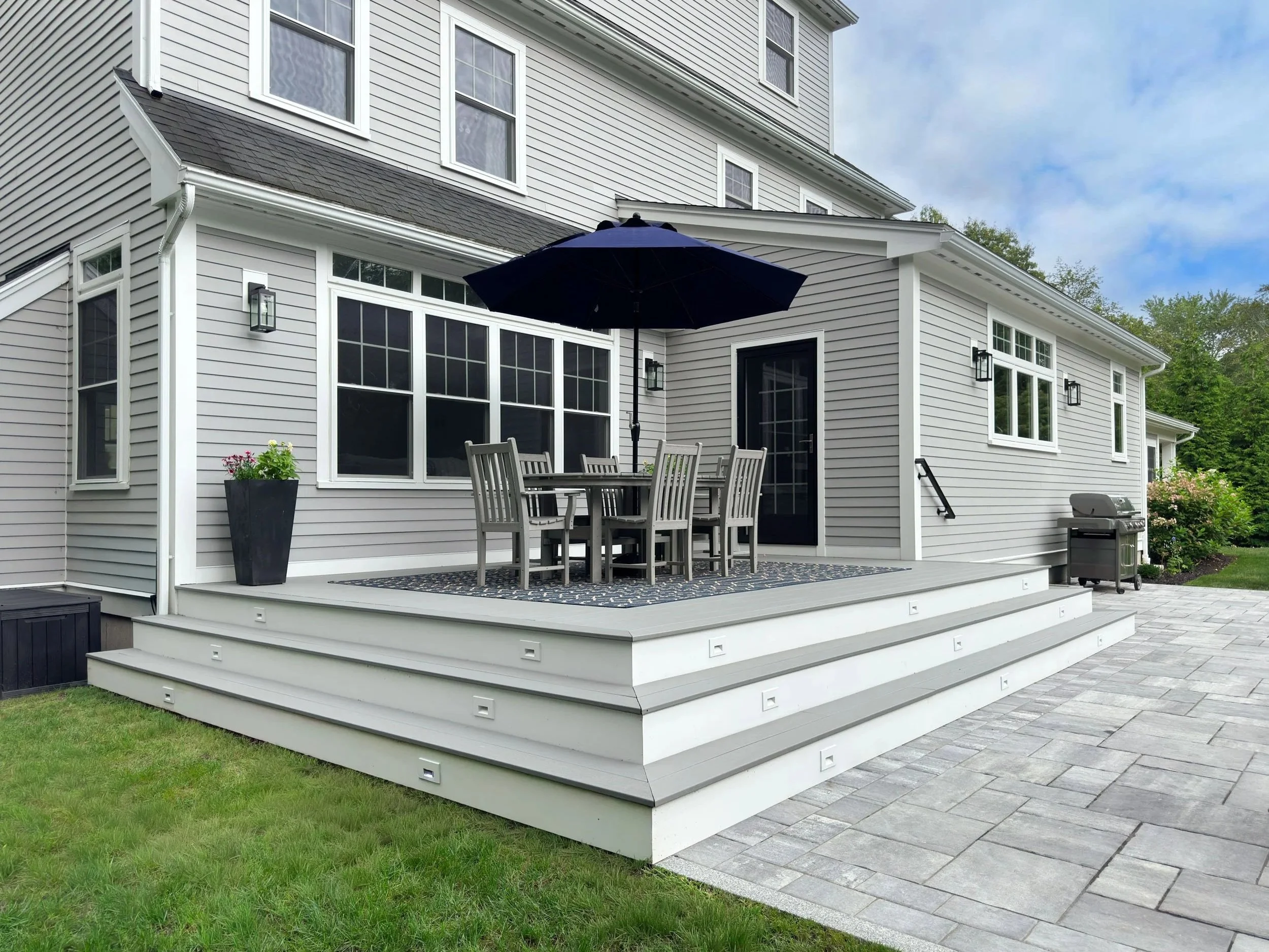 Timber Tech deck with gray dining set and navy umbrella overlooking patio below with three steps spilling out to the gathering spaces 