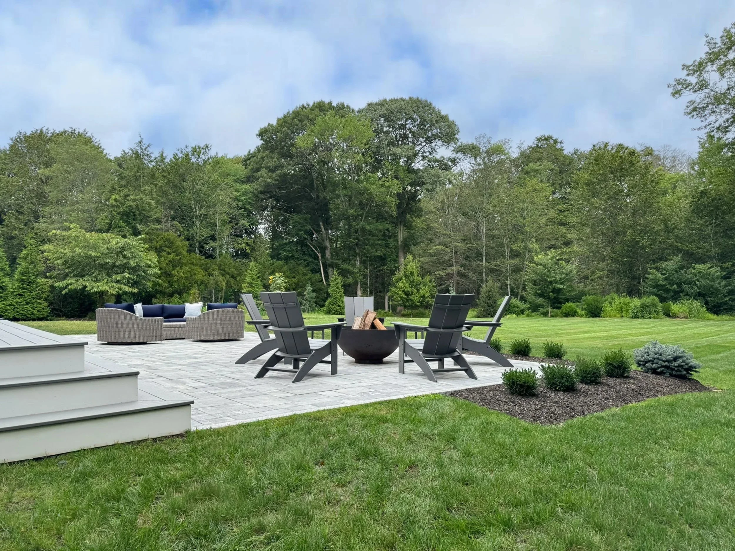 A firepit with Adirondack chairs and a lounge seating area beyond looking out into the yard at a vast wooded landscape.