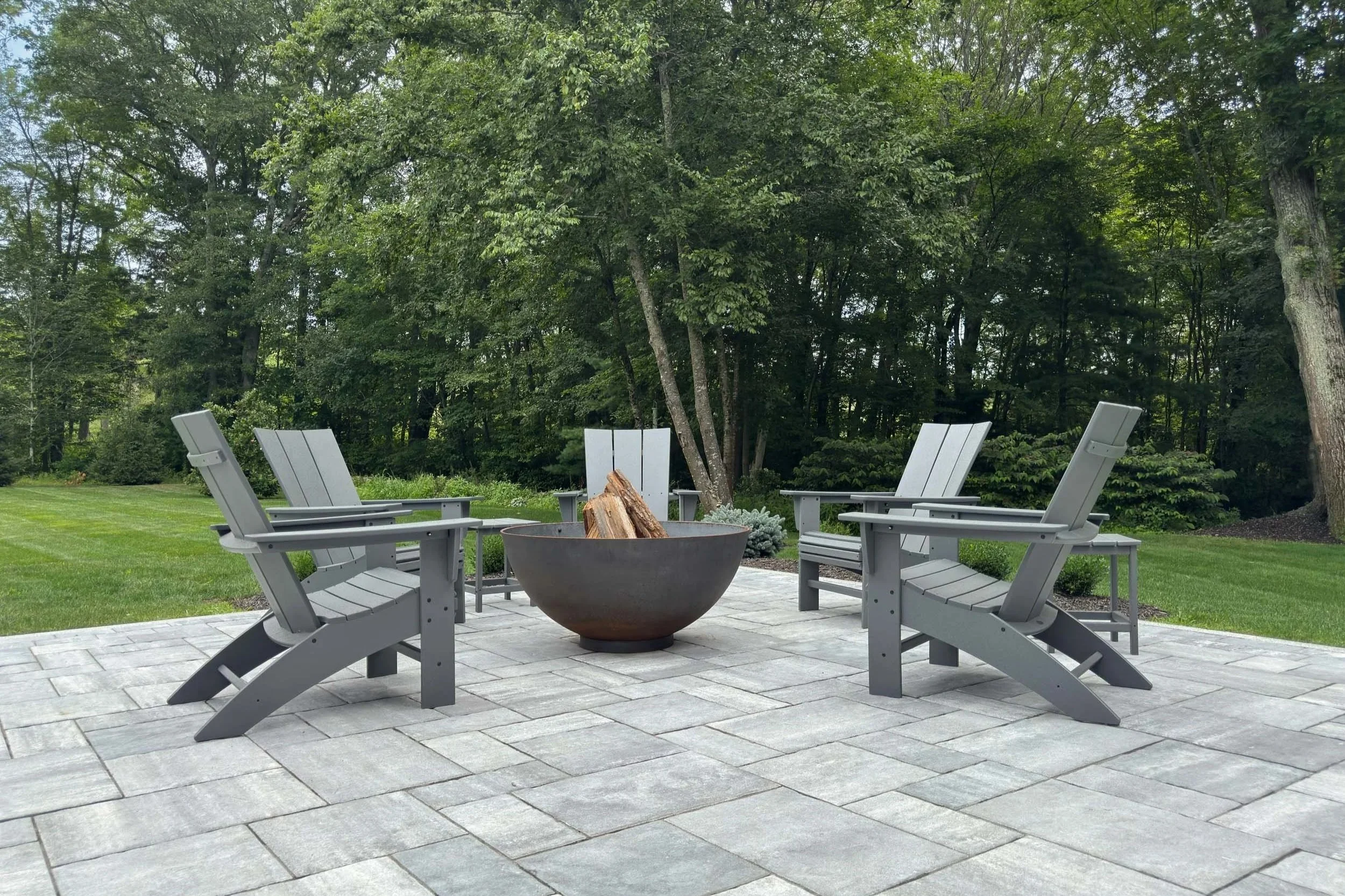 Polywood Adirondack chairs surrounding a corten steel bowl style fire pit on a large patio overlooking a densely wooded yard