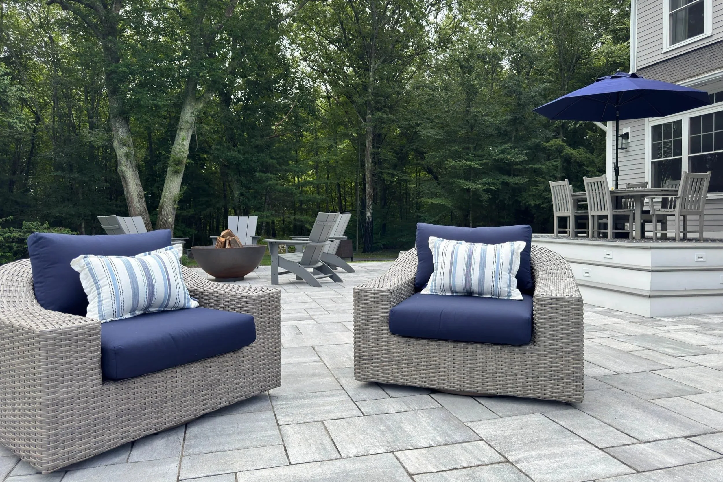 Two overside outdoor swivel chairs with navy cushions picture on a large grey concrete paver patio with fire pit and deck beyond.