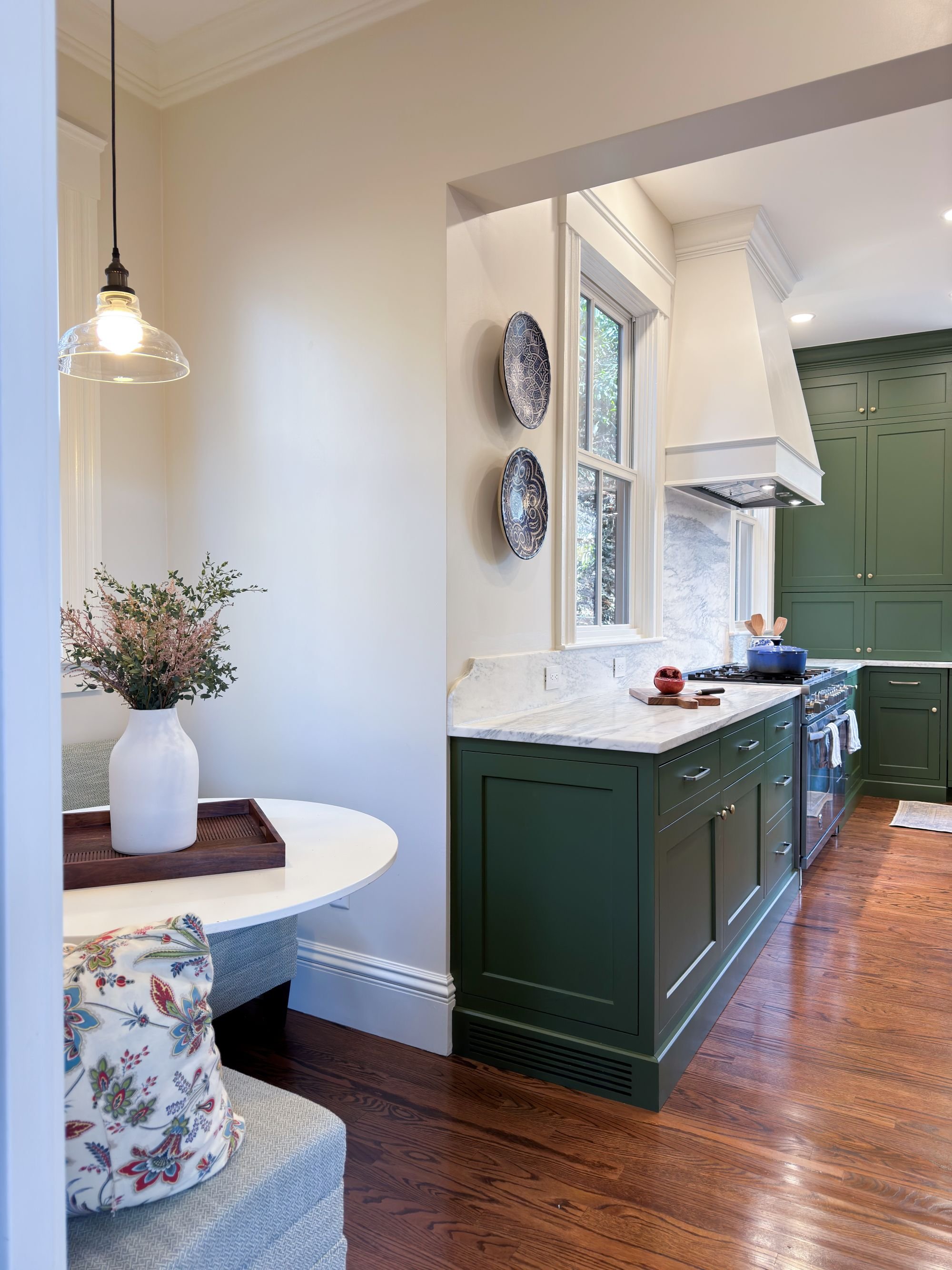 Renovated breakfast nook with view into kitchen of an 1846 Victorian home in Newton MA featuring banquette seating, green cabinetry and white range hood beyond
