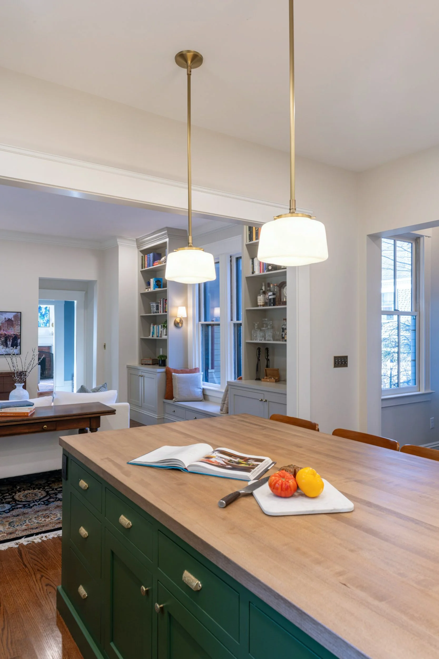 Renovated kitchen in an 1846 Victorian home in Newton MA showing open connection from butcher block island through to den with built-in bookshelves and brass pendant lighting