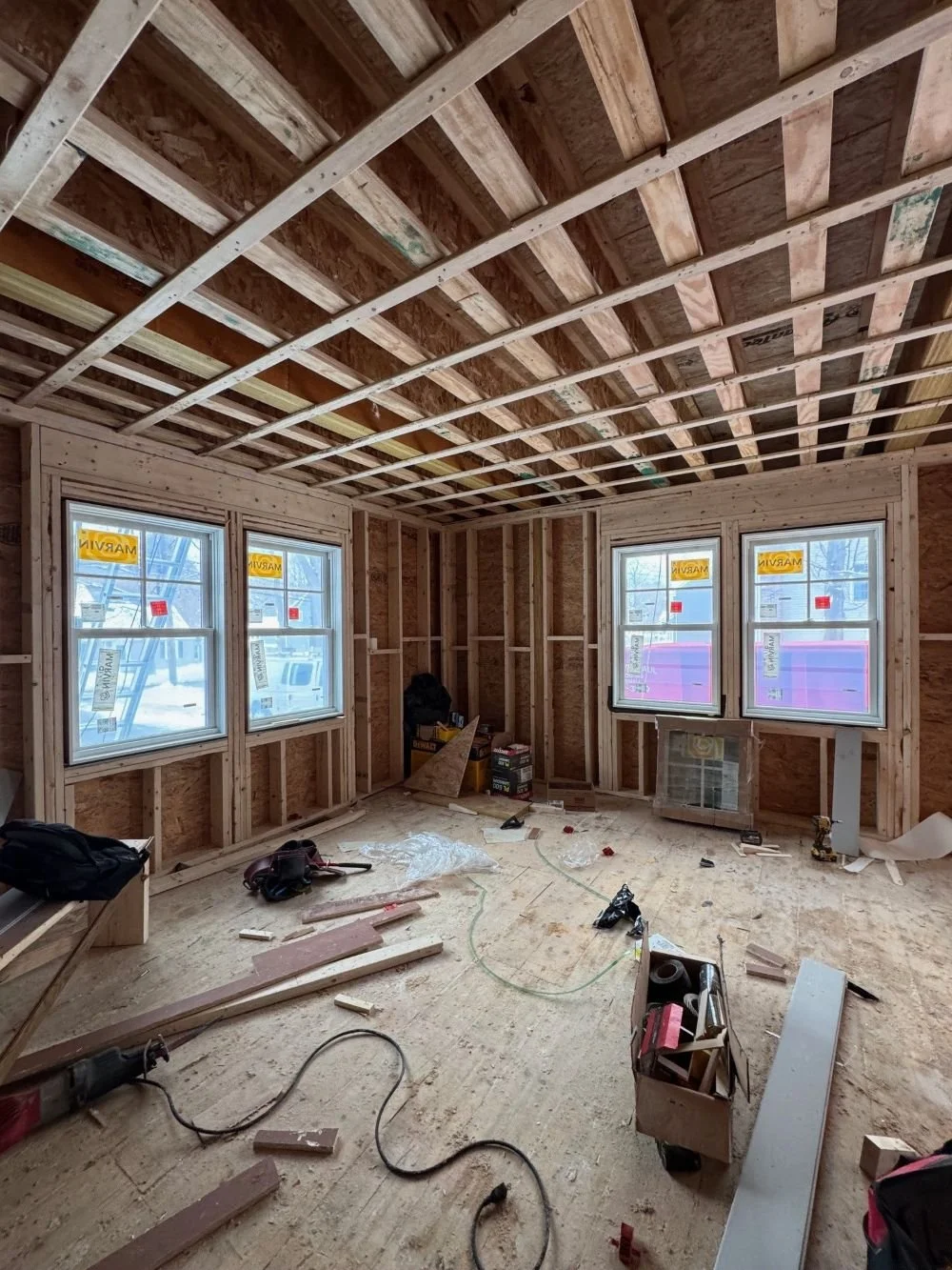 Ground level interior framing phase of a Dutch Colonial second story addition in Greater Boston showing new window openings and structural framing designed by Tact and Grit Design Co.