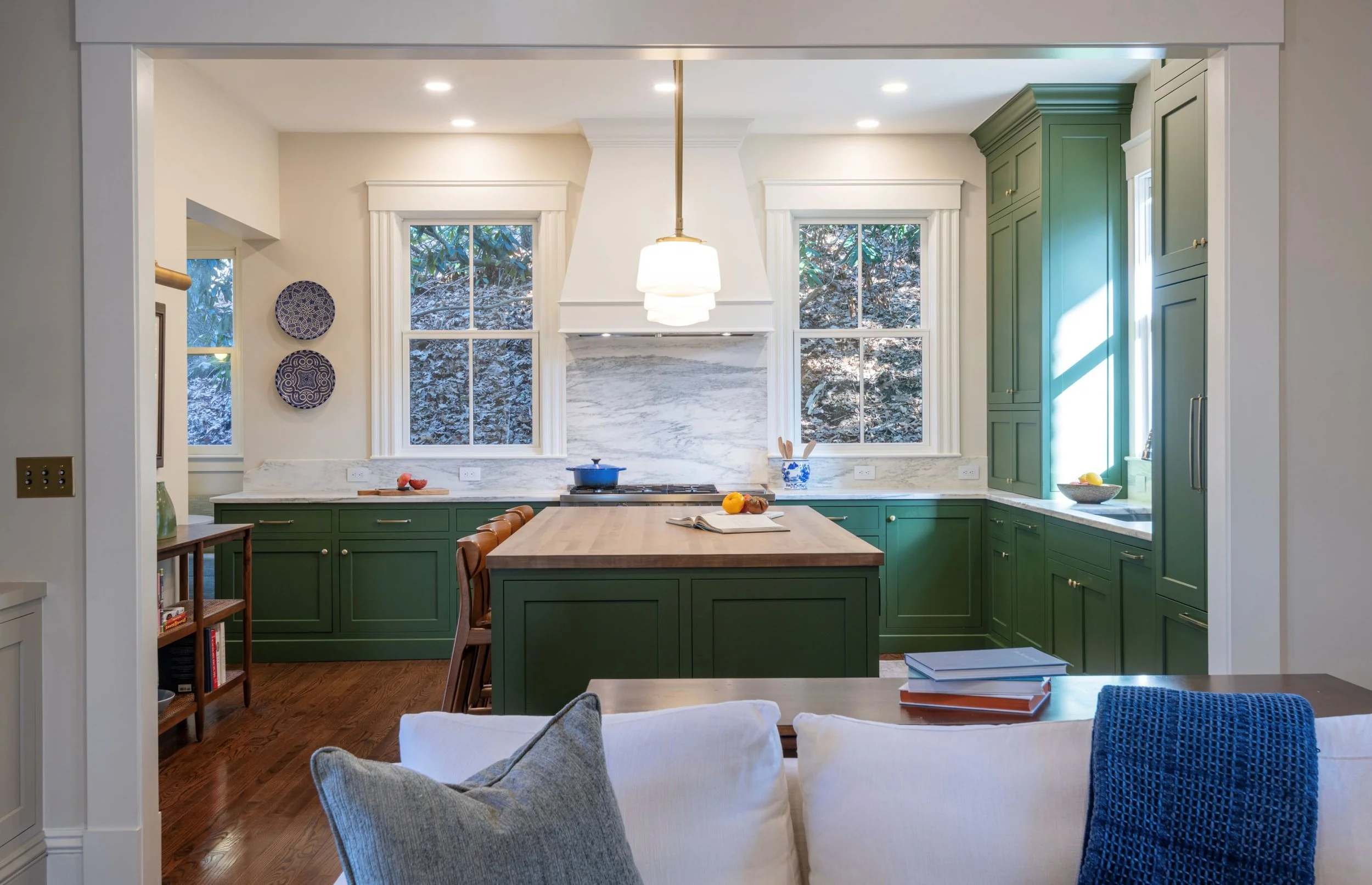 Renovated kitchen in an 1846 Victorian home in Newton MA featuring custom green cabinetry, butcher block island with seating, marble backsplash and white range hood with view from living room