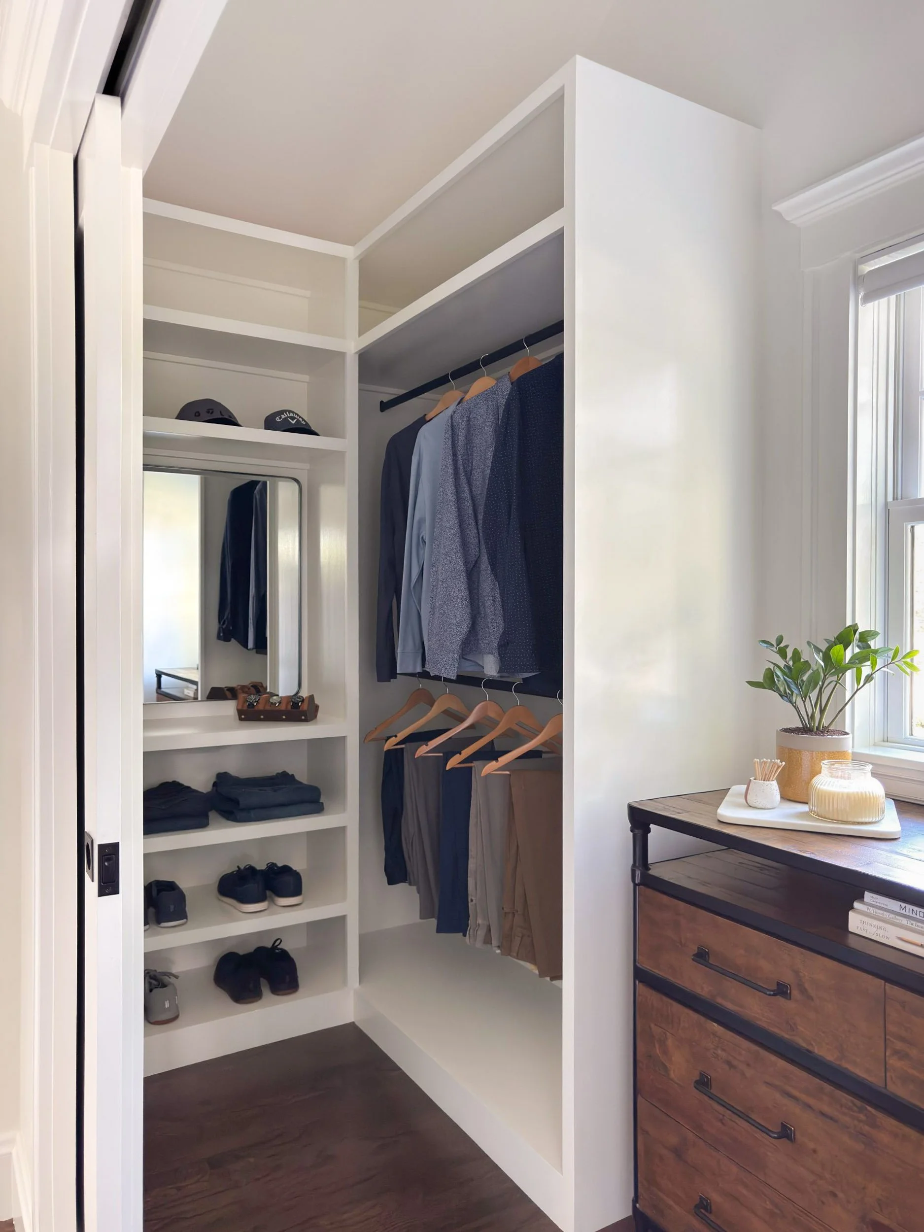 Completed walk-in closet of a Dutch Colonial primary suite addition in Greater Boston showing custom built-in shelving, hanging rods and reclaimed wood and iron dresser under window.