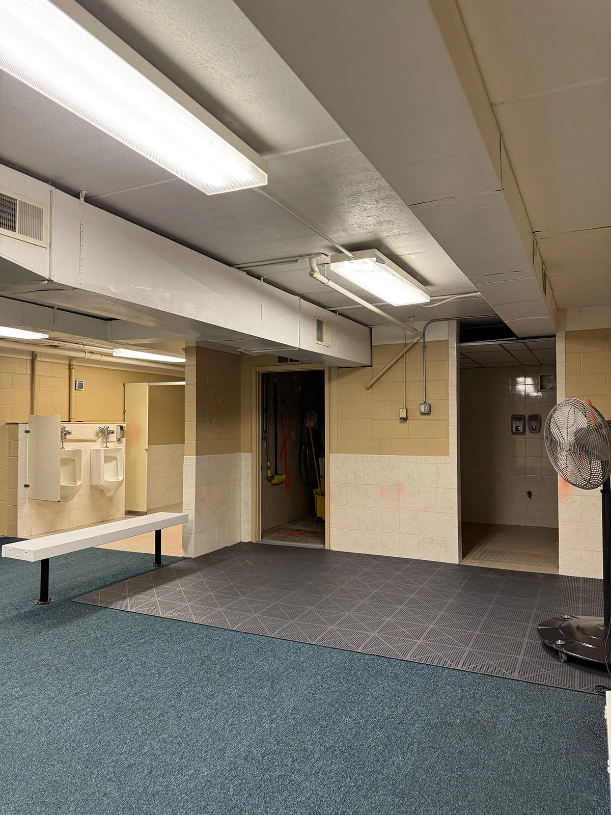 Original view of a dated commercial locker room featuring exposed mechanicals, concrete block walls, and industrial lighting prior to a full architectural reconfiguration."