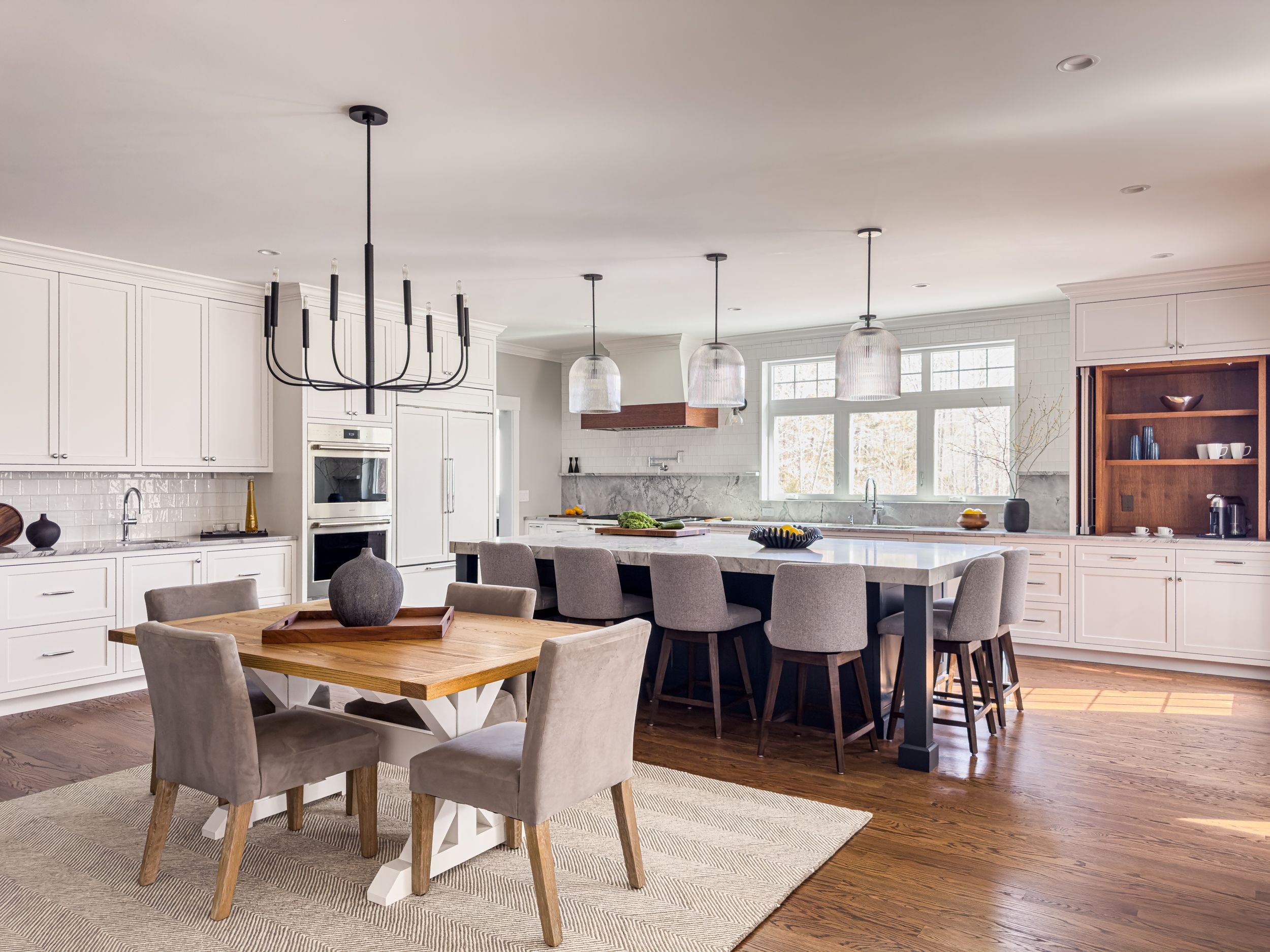 Open-concept casual dining and kitchen with black chandelier, fluted glass pendants, large dark island, custom inset white cabinetry, and stained white oak accent cabinet in Hamilton MA home by Tact & Grit Design Co.