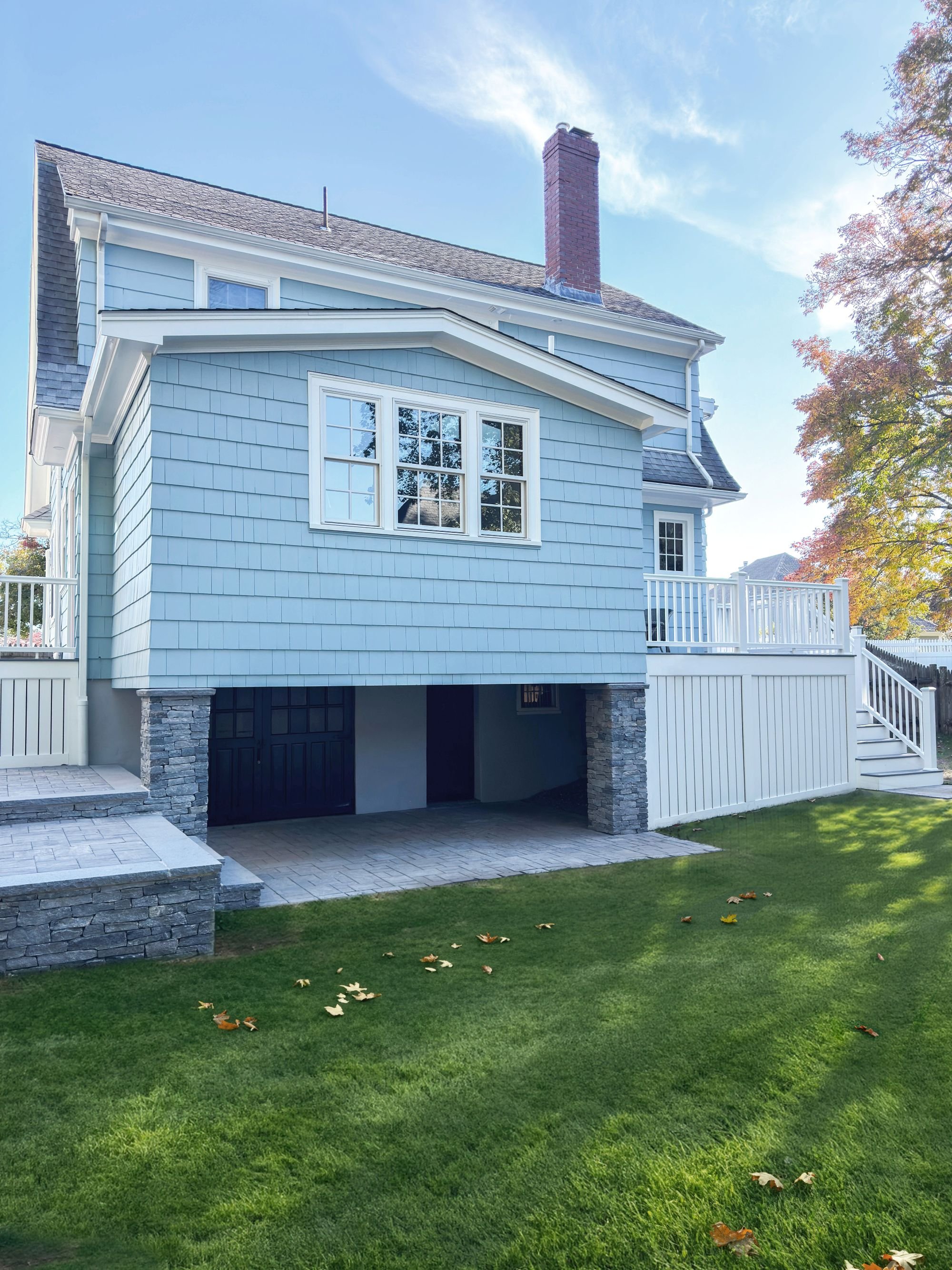 Renovated kitchen addition exterior with light blue cedar shake siding, triple window, stone piers, and open below grade storage, set against original Dutch Colonial home in Medford MA