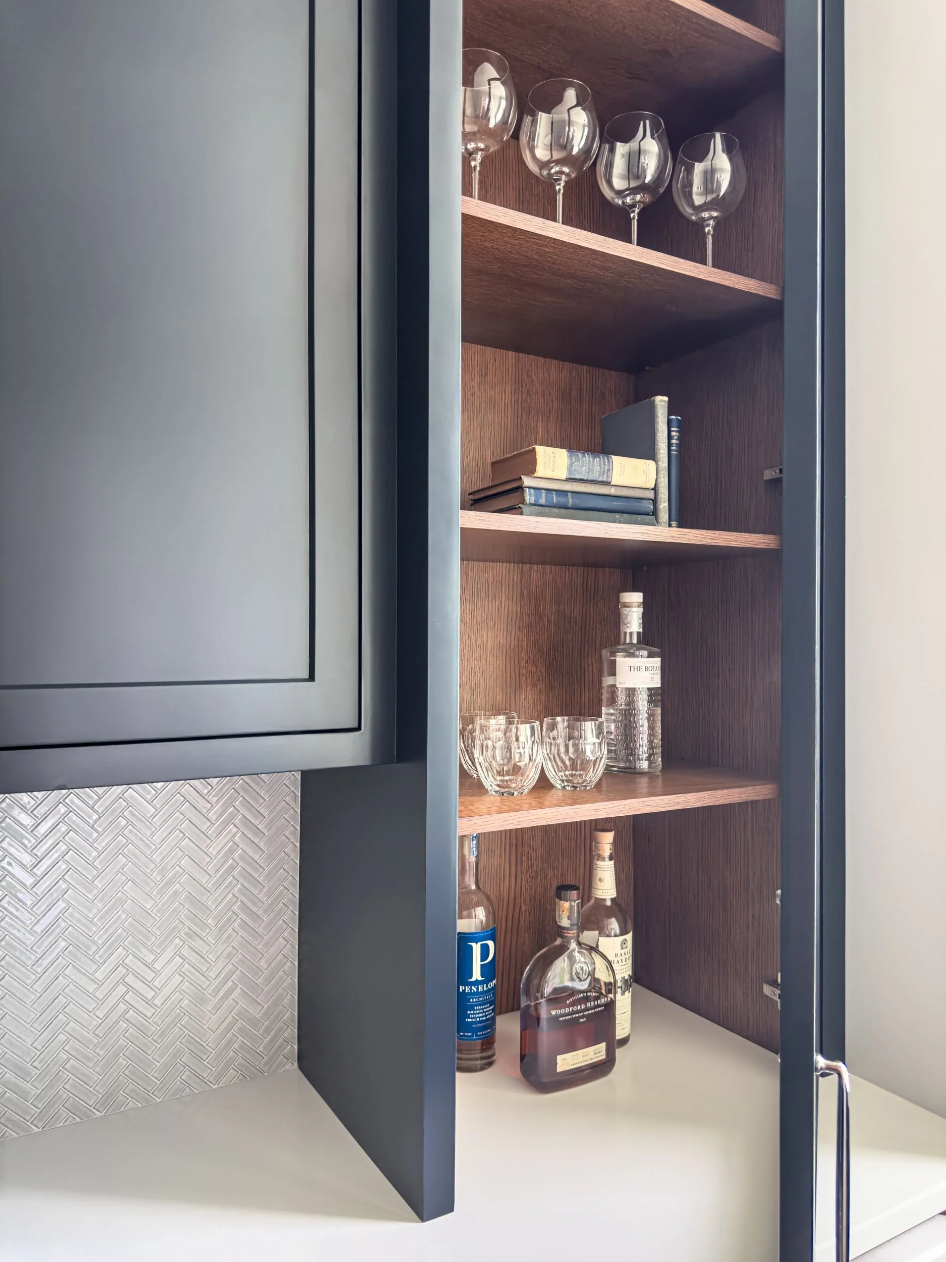 Custom wine glass cabinet with stained white oak interior, bar storage, herringbone tile backsplash, and navy counter to ceiling accent cabinet in Hamilton MA butler's pantry by Tact & Grit Design Co.