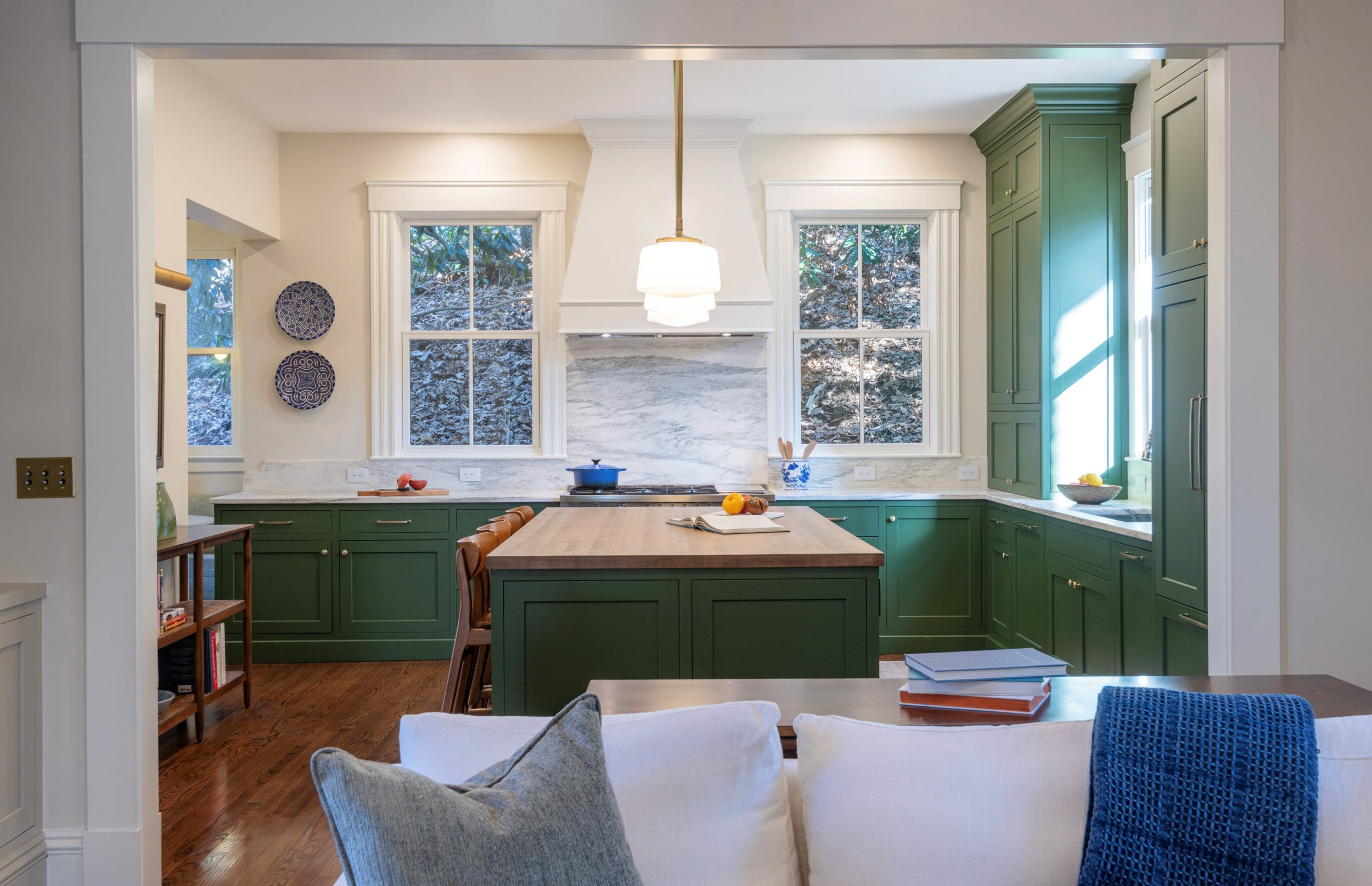 Renovated kitchen in an 1846 Victorian home in Newton MA featuring custom green cabinetry, butcher block island with seating, marble backsplash and white range hood with view from living room