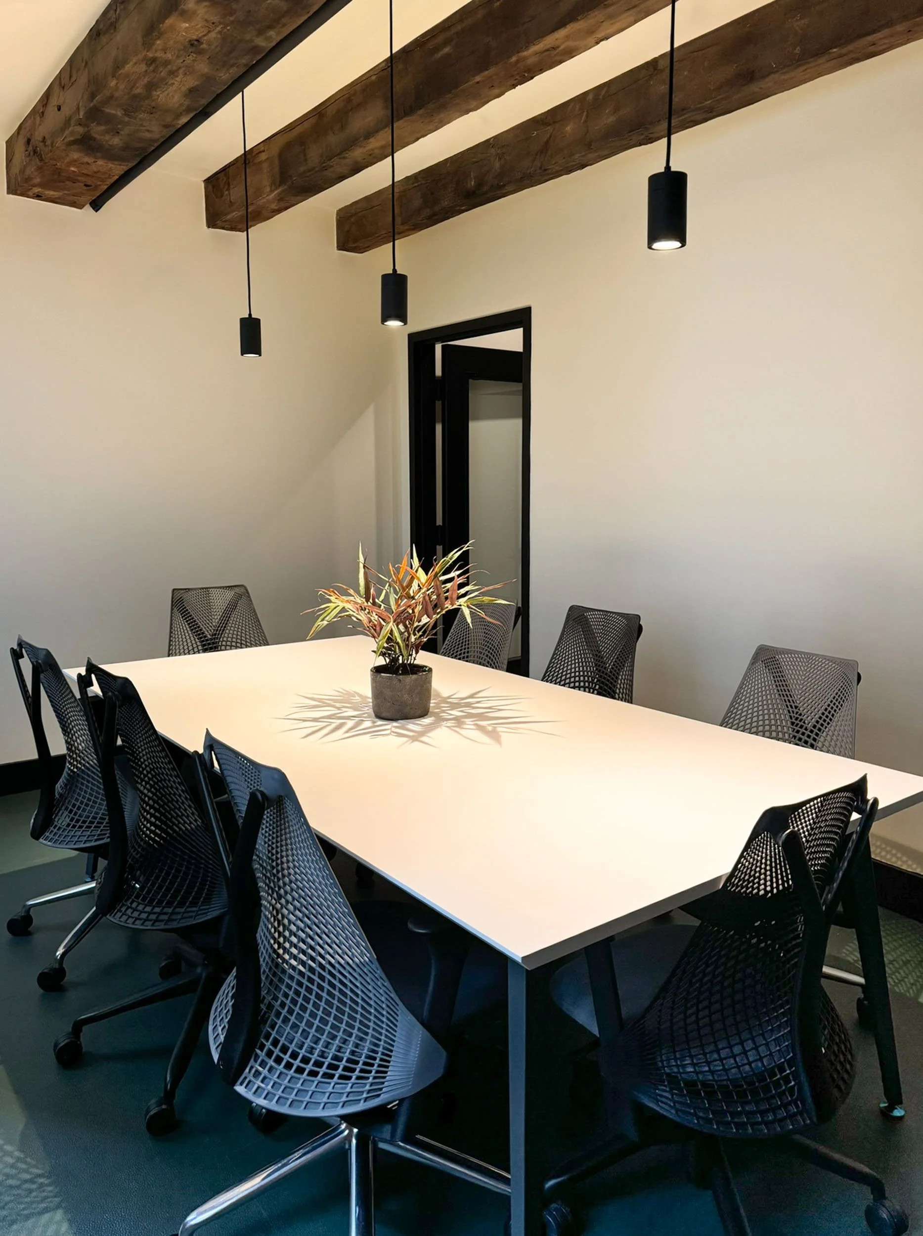 A conference room with a long white table, mesh-back chairs and pendant lighting beneath original wood beams.