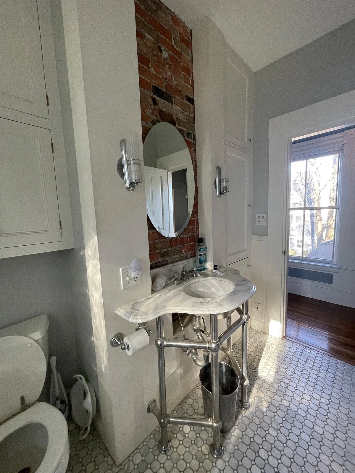 Alt text: Existing guest bathroom of an 1846 Victorian home in Newton MA prior to reconfiguration showing pedestal sink, exposed brick chimney and original marble hex tile floor