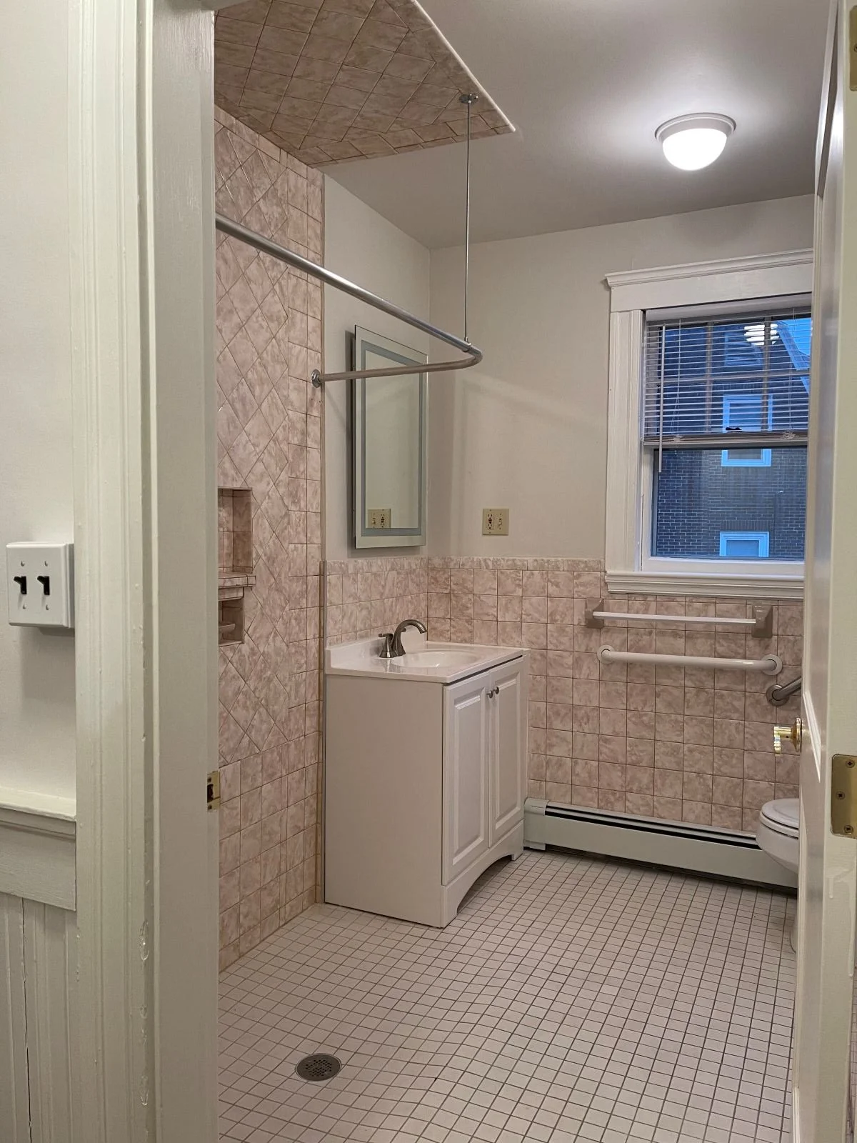 View of existing antique pink tiled bathroom from entry way picturing partial shower and vanity.
