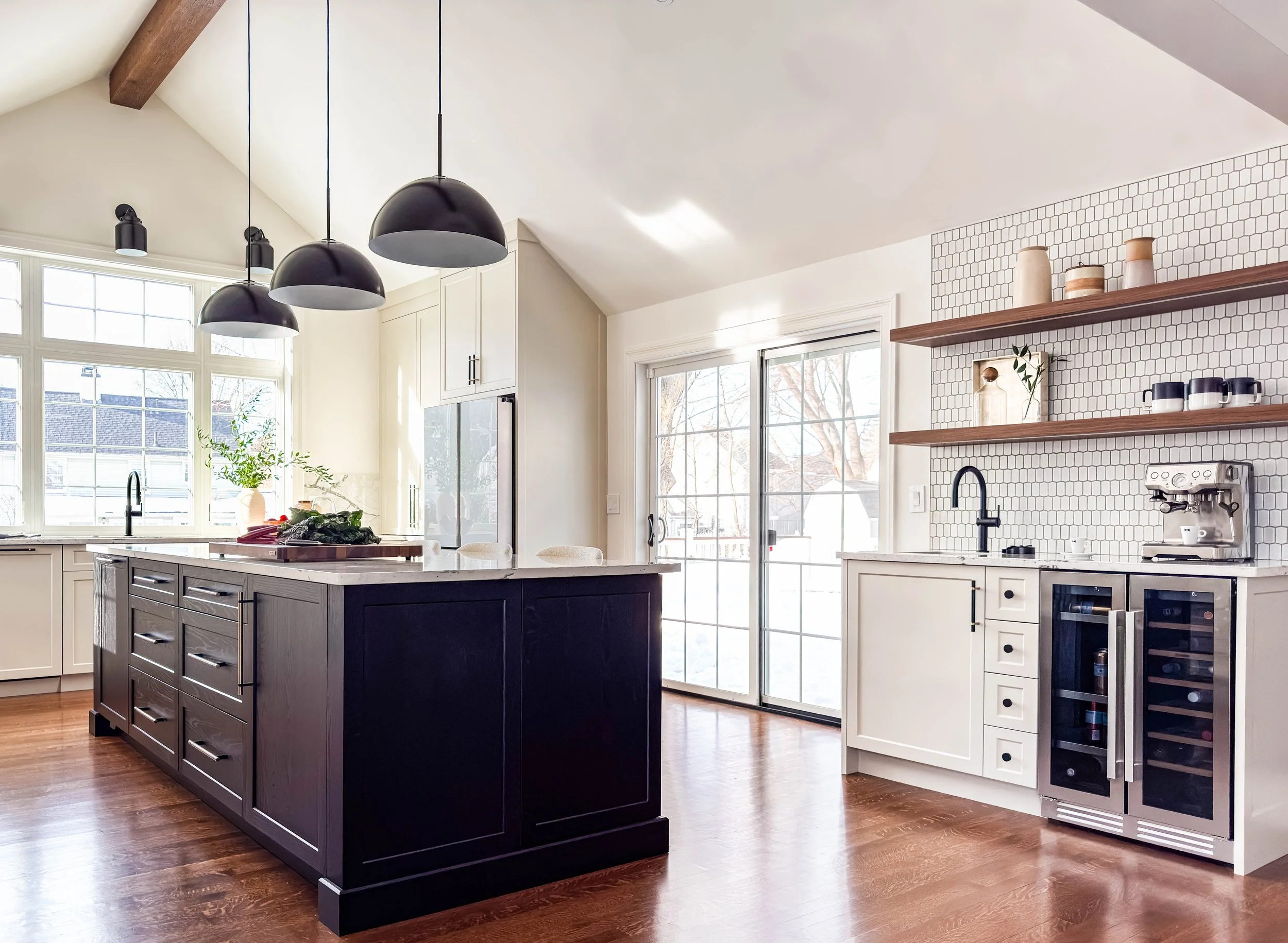 Kitchen addition featuring a dedicated coffee bar and beverage station with open wood shelving, a wine fridge, and a picket tile backsplash. A large stained black oak island sits under a vaulted ceiling, adjacent to sliding glass doors that open onto