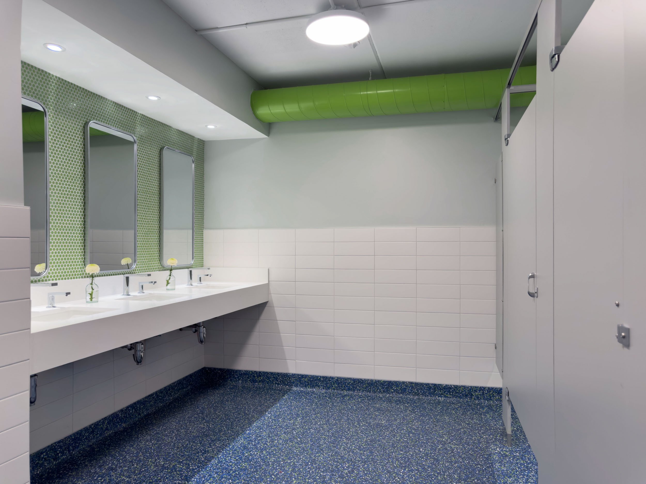 women's locker room sink and toilet area with three sinks and a green penny backsplash and grey stalls with blue and green epoxy floor