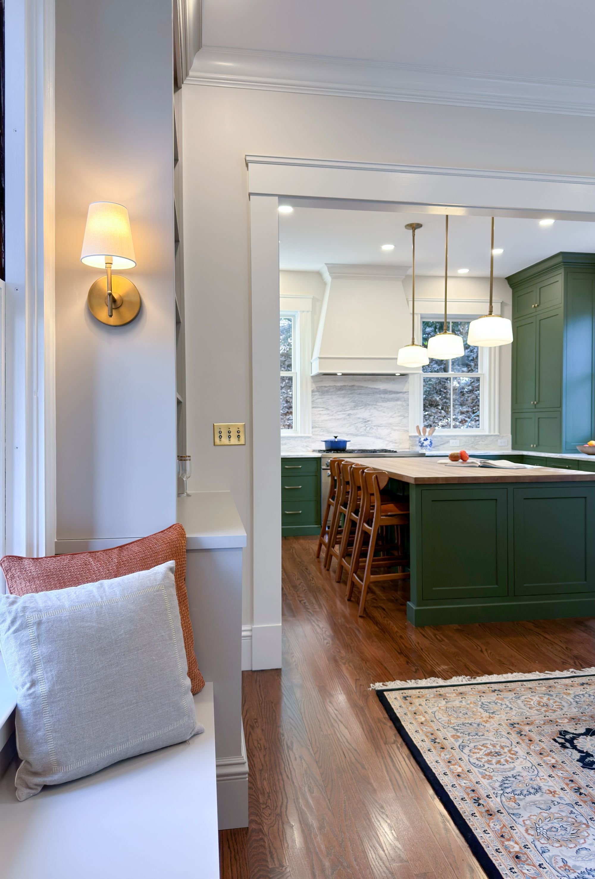 View from new den built-ins featuring a reading bench into the kitchen of an 1846 Victorian home in Newton MA with green cabinetry beyond