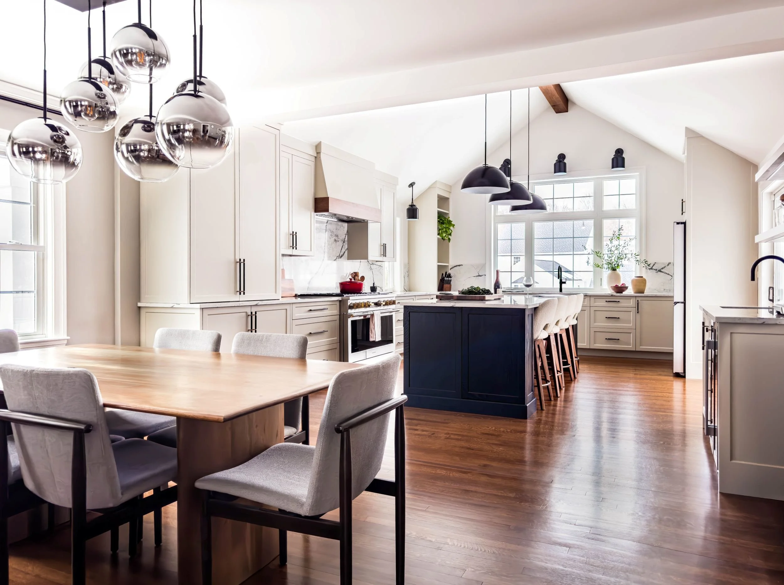 Open-concept dining and kitchen addition featuring a vaulted ceiling with exposed wood beam, a stained black oak statement island, and modern globe pendant lighting over a natural wood dining table.