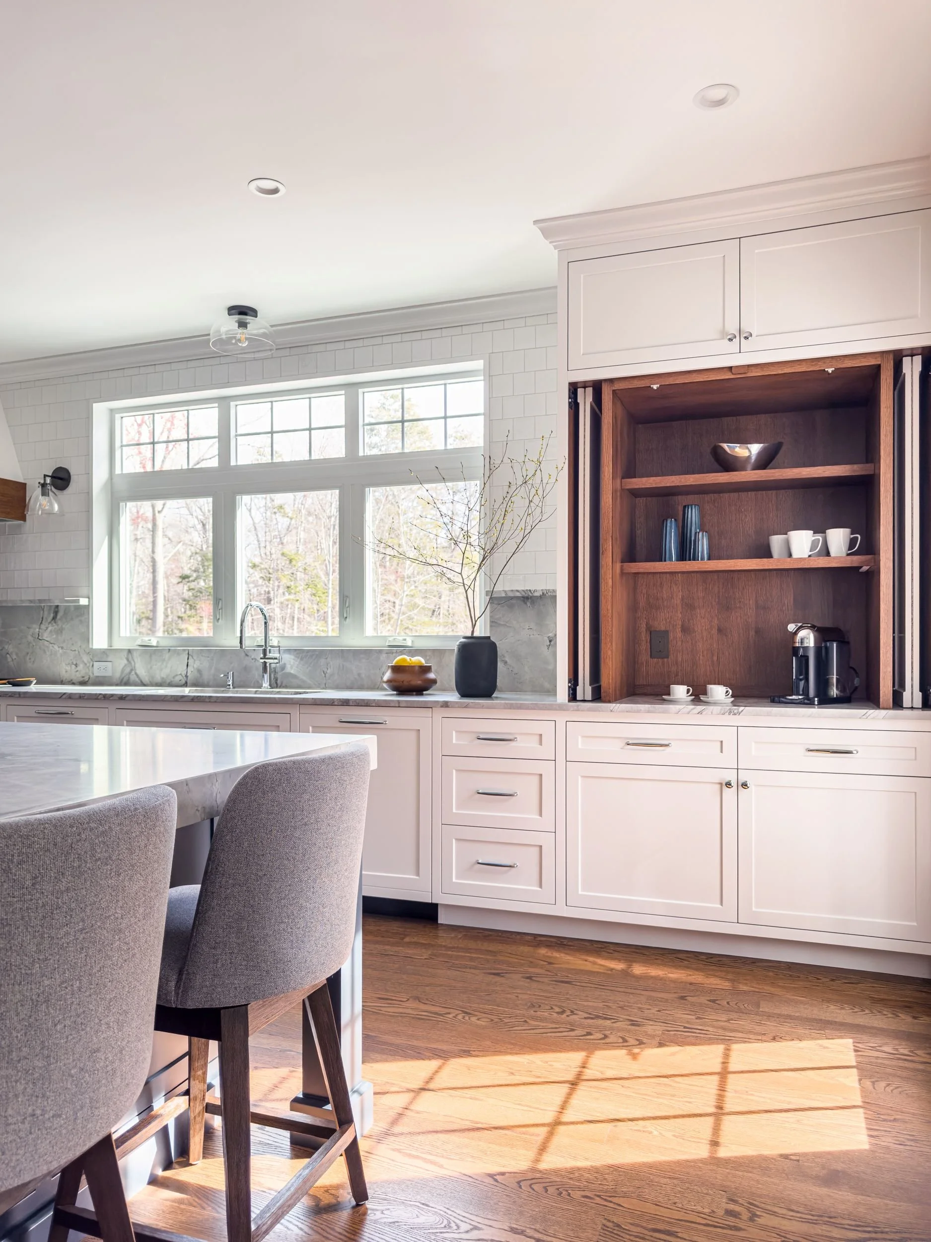 Kitchen island seating area with gray upholstered barstools, stained white oak accent coffee cabinet, and natural light windows in Hamilton MA home by Tact & Grit Design Co.